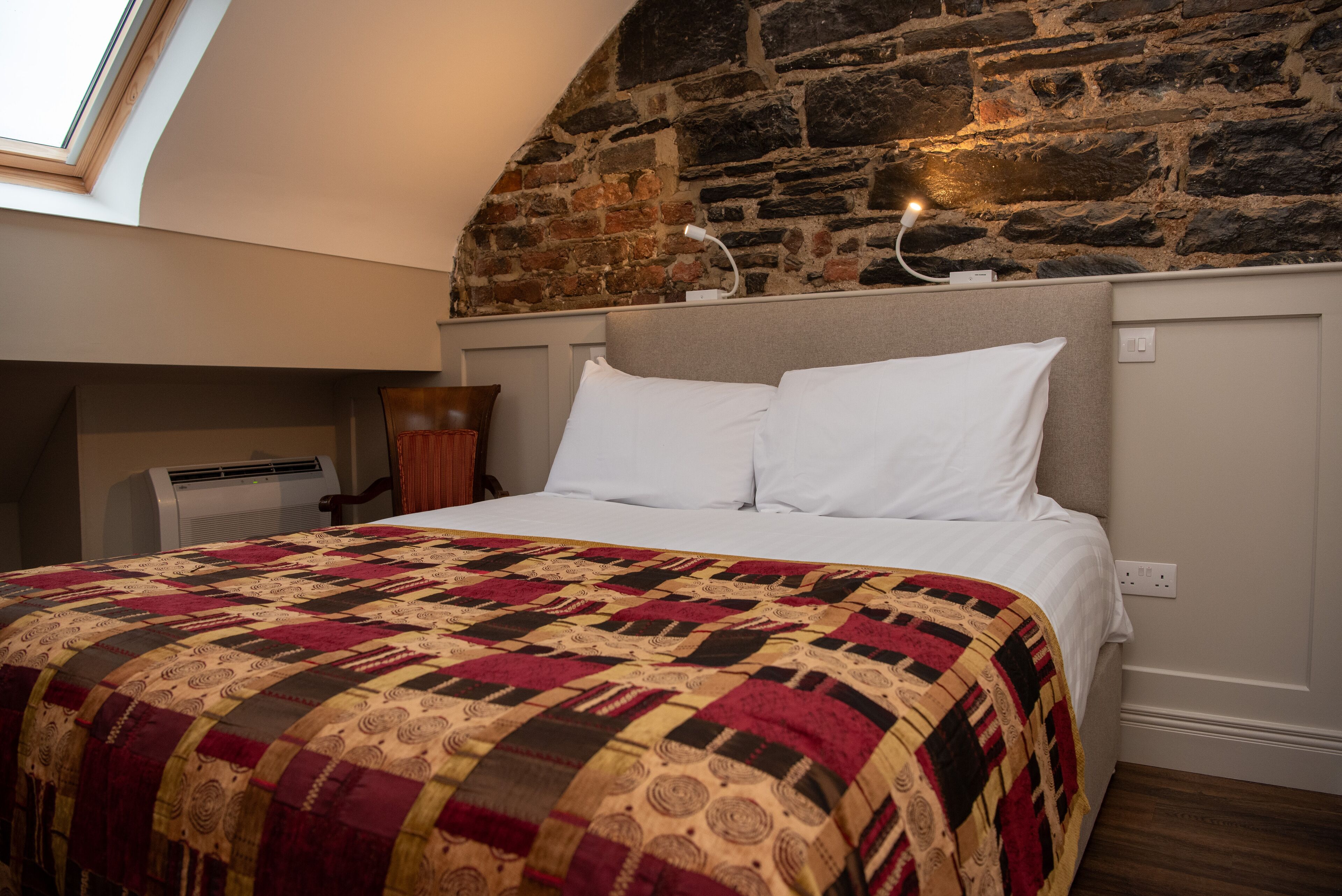 A bedroom with a sloped stone wall, a skylight, a bed with white linens, and a patterned quilt.