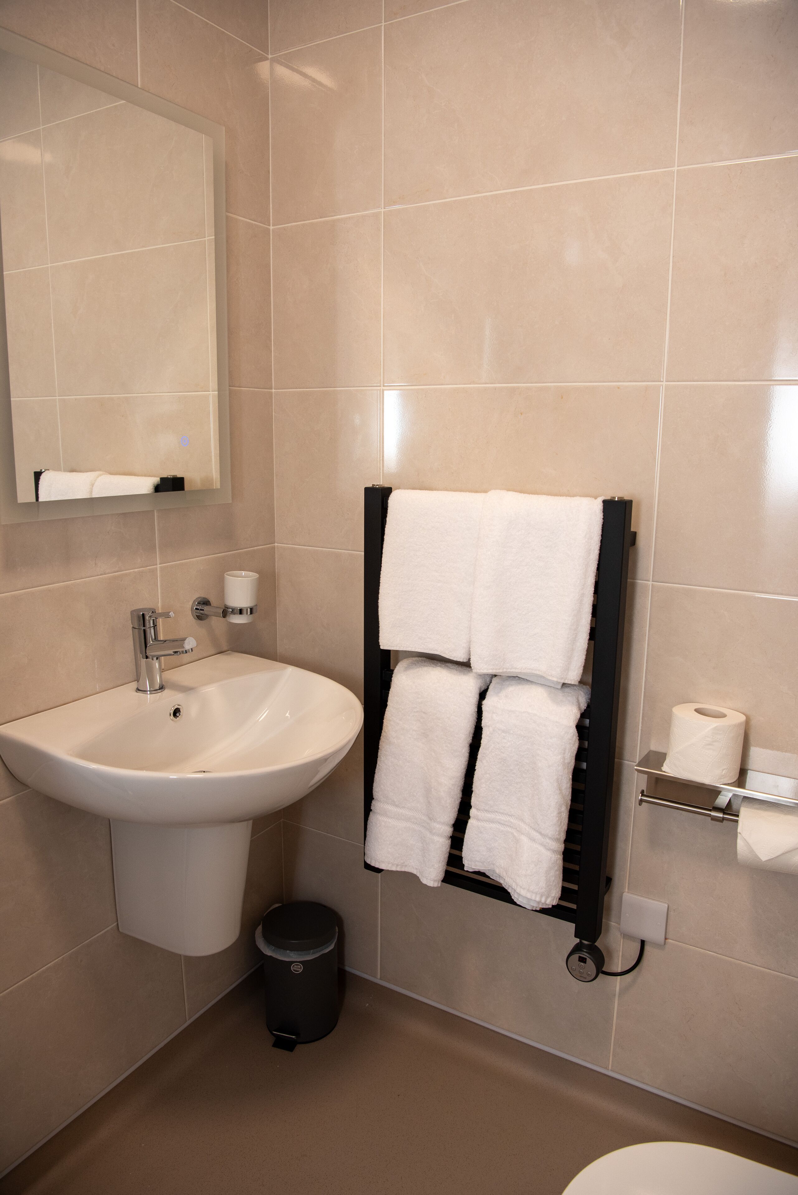 Un baño con un lavabo blanco, un espejo, un perchero con dos toallas, un cubo de basura negro y un dispensador de papel higiénico.