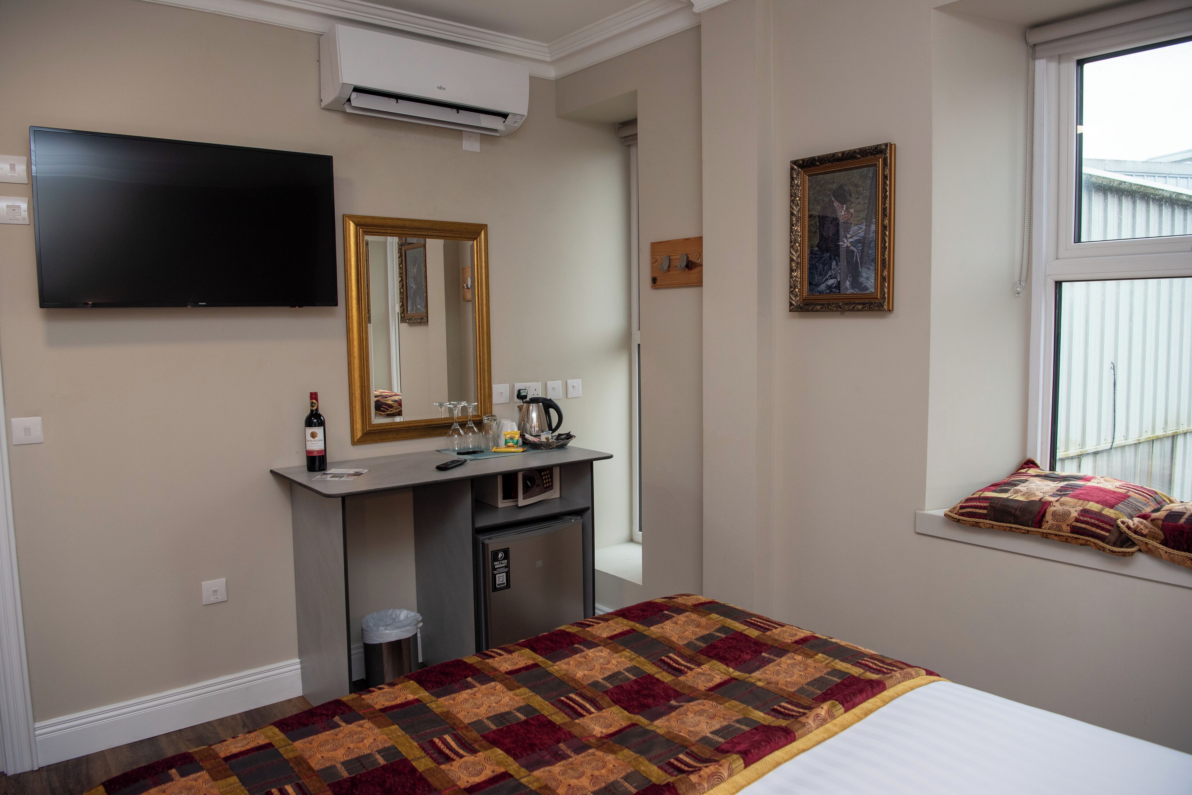 A hotel room with a bed, a desk with a mirror, a television, a window with a view, and a small refrigerator.