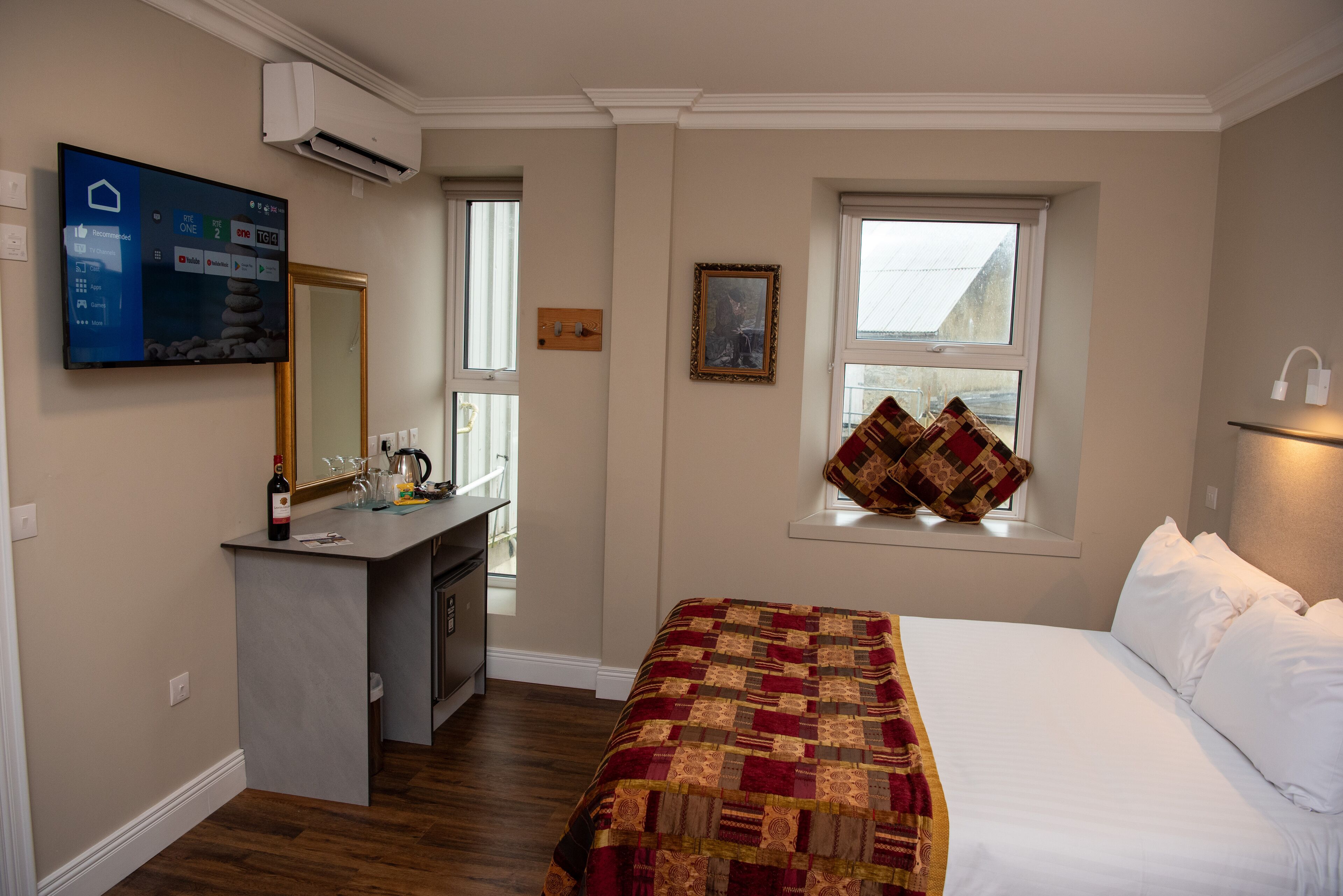 A hotel room with a bed, a desk with a mirror, a TV, a window with a view, and a painting on the wall.