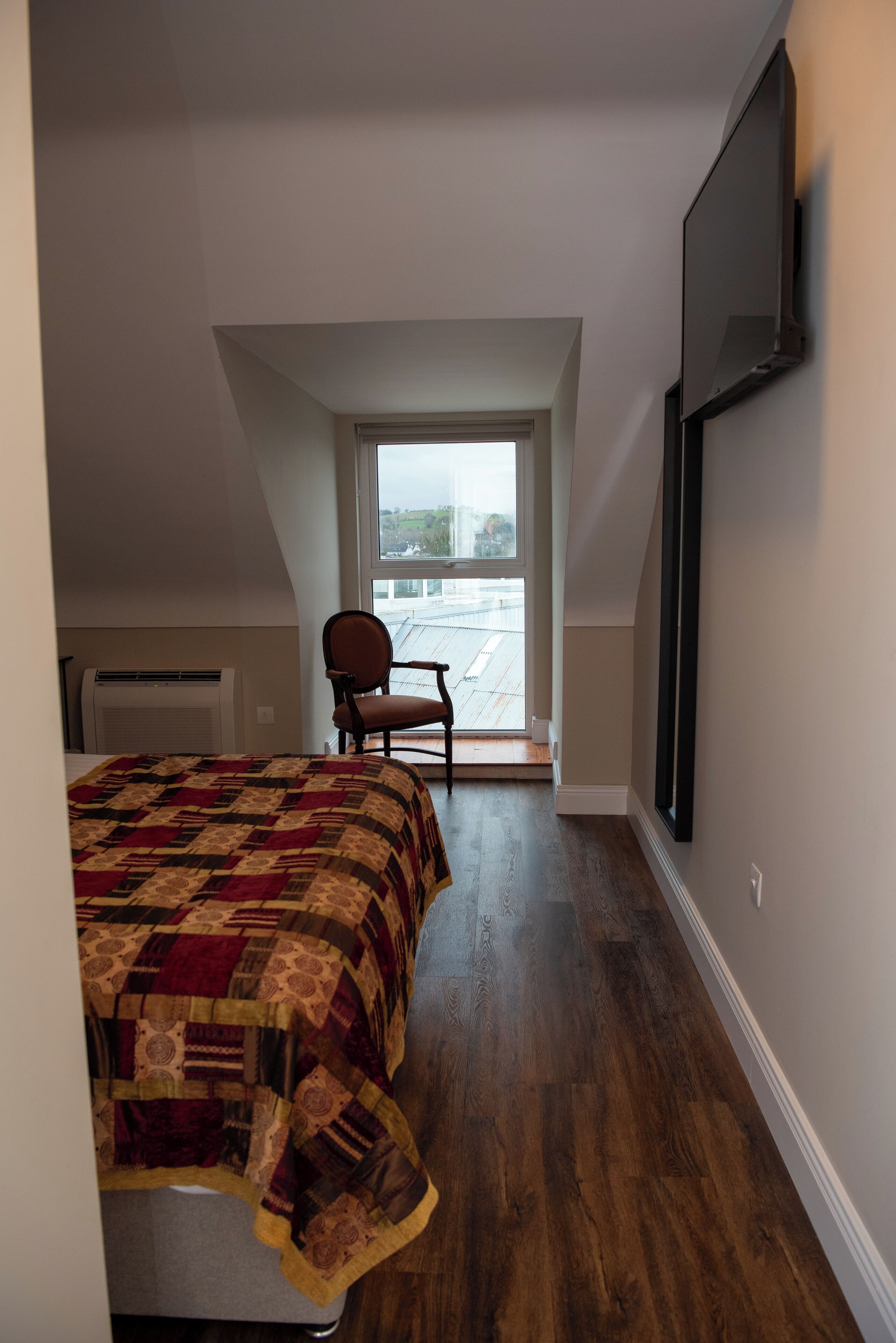 Un dormitorio con una cama grande, una silla, un televisor montado en la pared y un ventanal con vistas al exterior.
