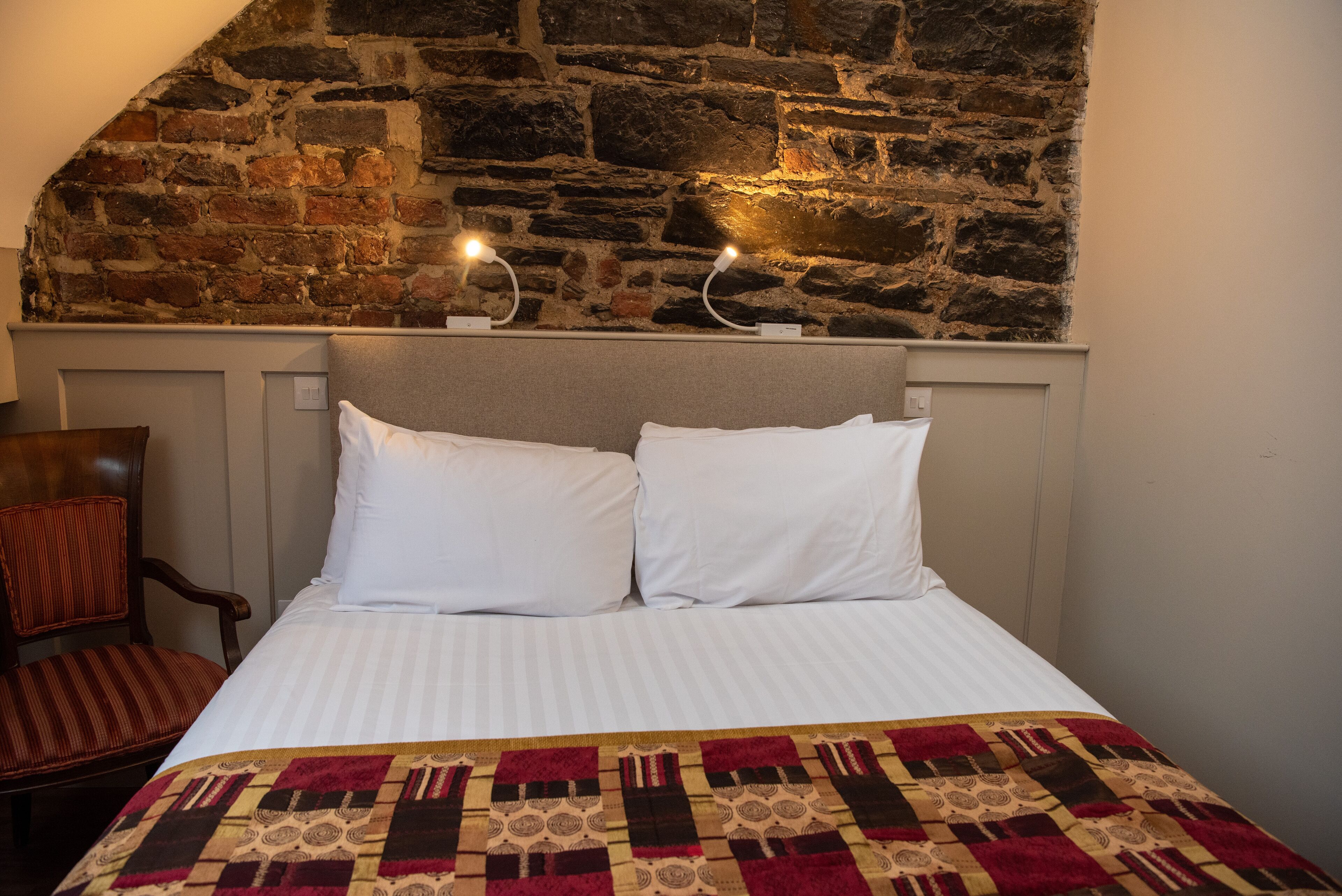 Un dormitorio con pared de piedra, cama con ropa de cama blanca, manta con estampado y una silla.