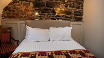 Un dormitorio con pared de piedra, cama con ropa de cama blanca, manta con estampado y una silla.