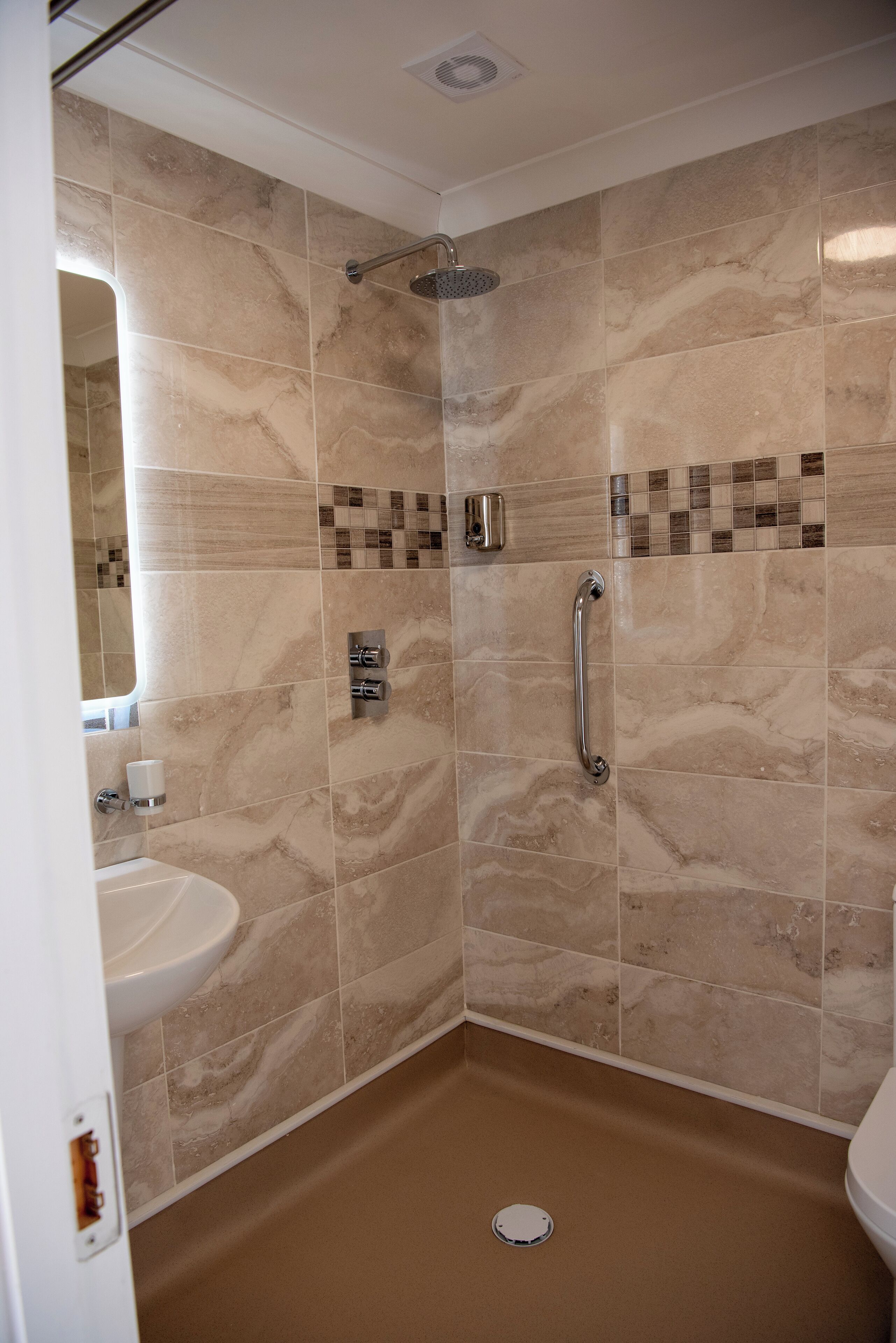 A bathroom with beige marble tiles, a shower with a handheld showerhead, a toilet, and a sink with a mirror.