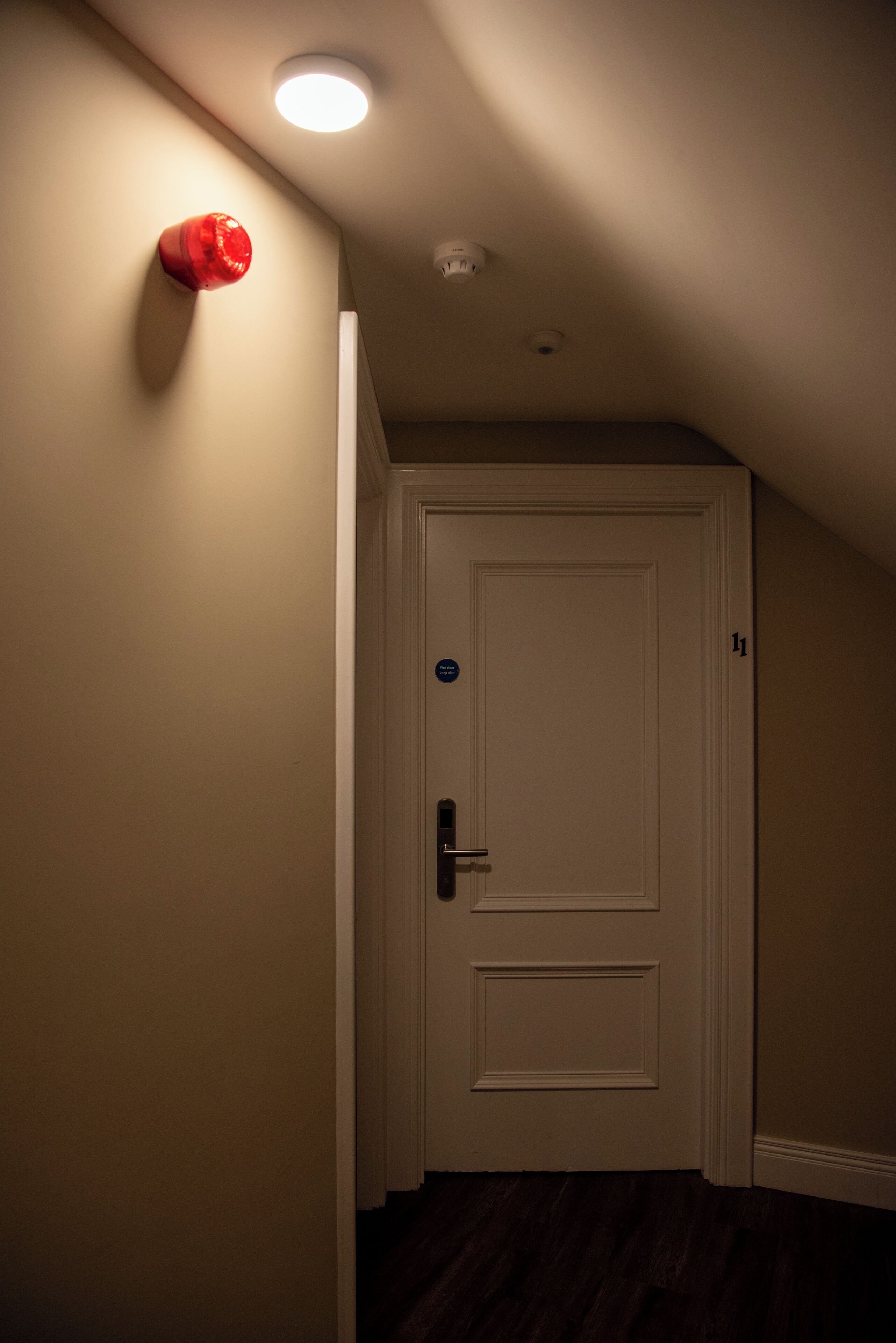 A narrow hallway with a white door, a red emergency light, and a ceiling light.