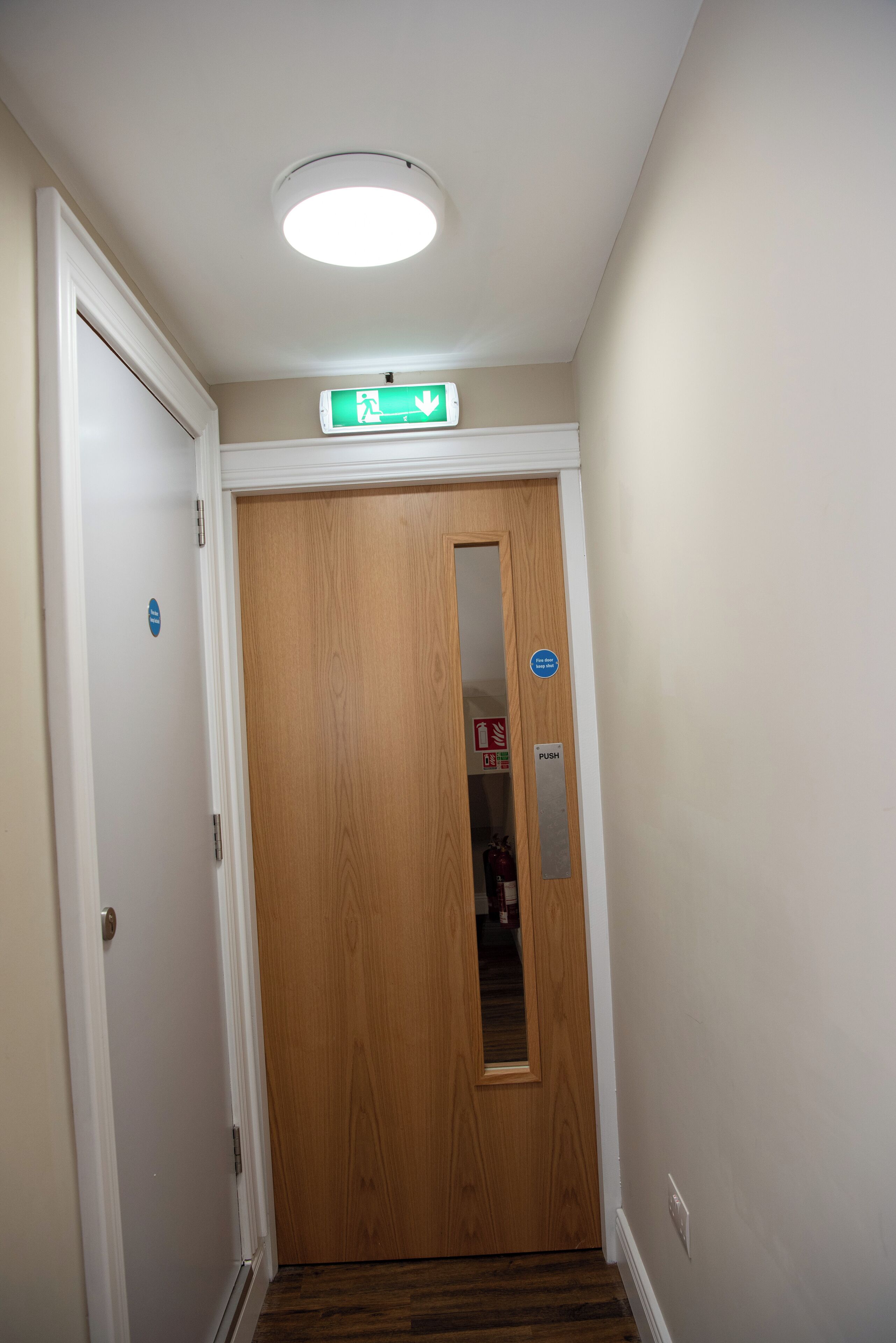 Un couloir avec une porte en bois, une porte blanche et un luminaire au plafond.