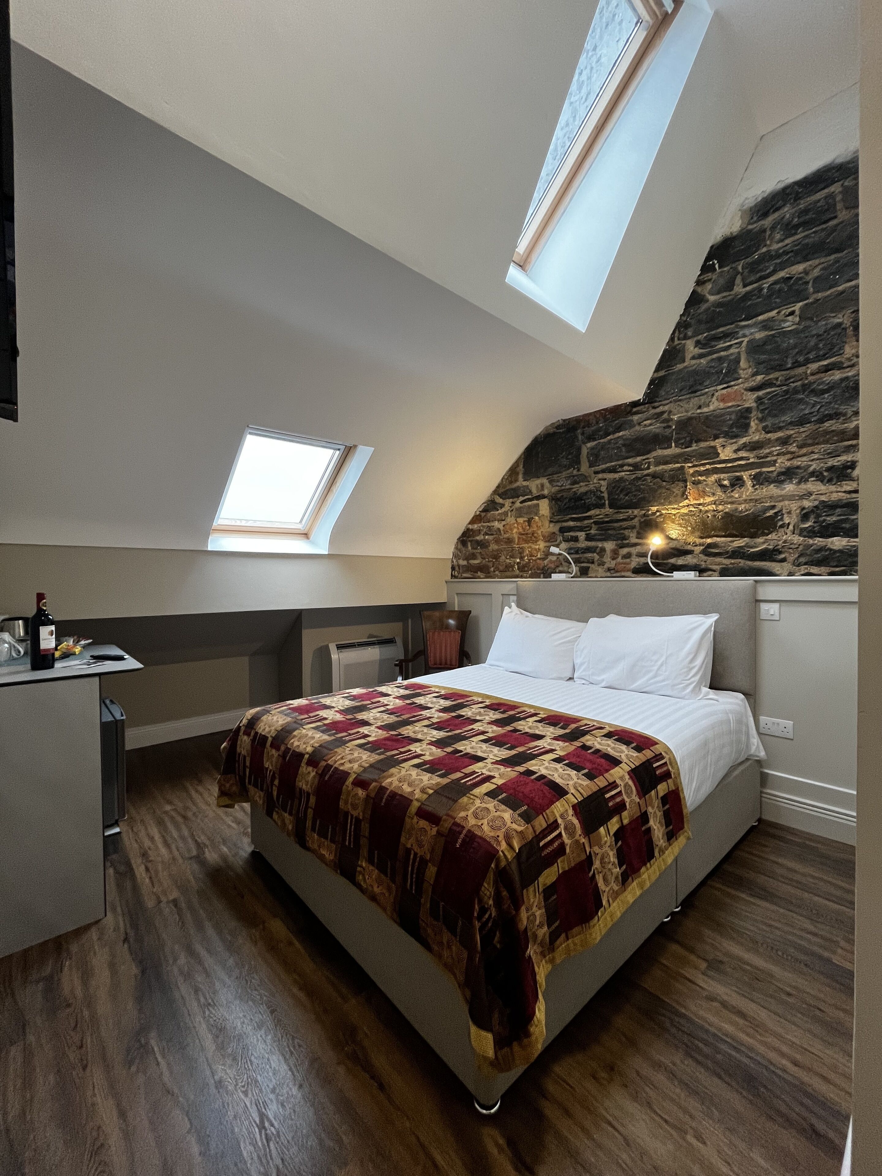 Een slaapkamer met een schuine kap, een stenen muur, een bed met een geborduurd kleed, een bureau met een fles en een stoel.