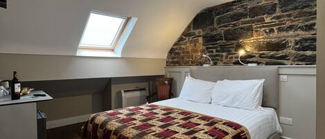 A bedroom with a sloped ceiling, a stone wall, a bed with a patterned blanket, a desk with a bottle, and a chair.
