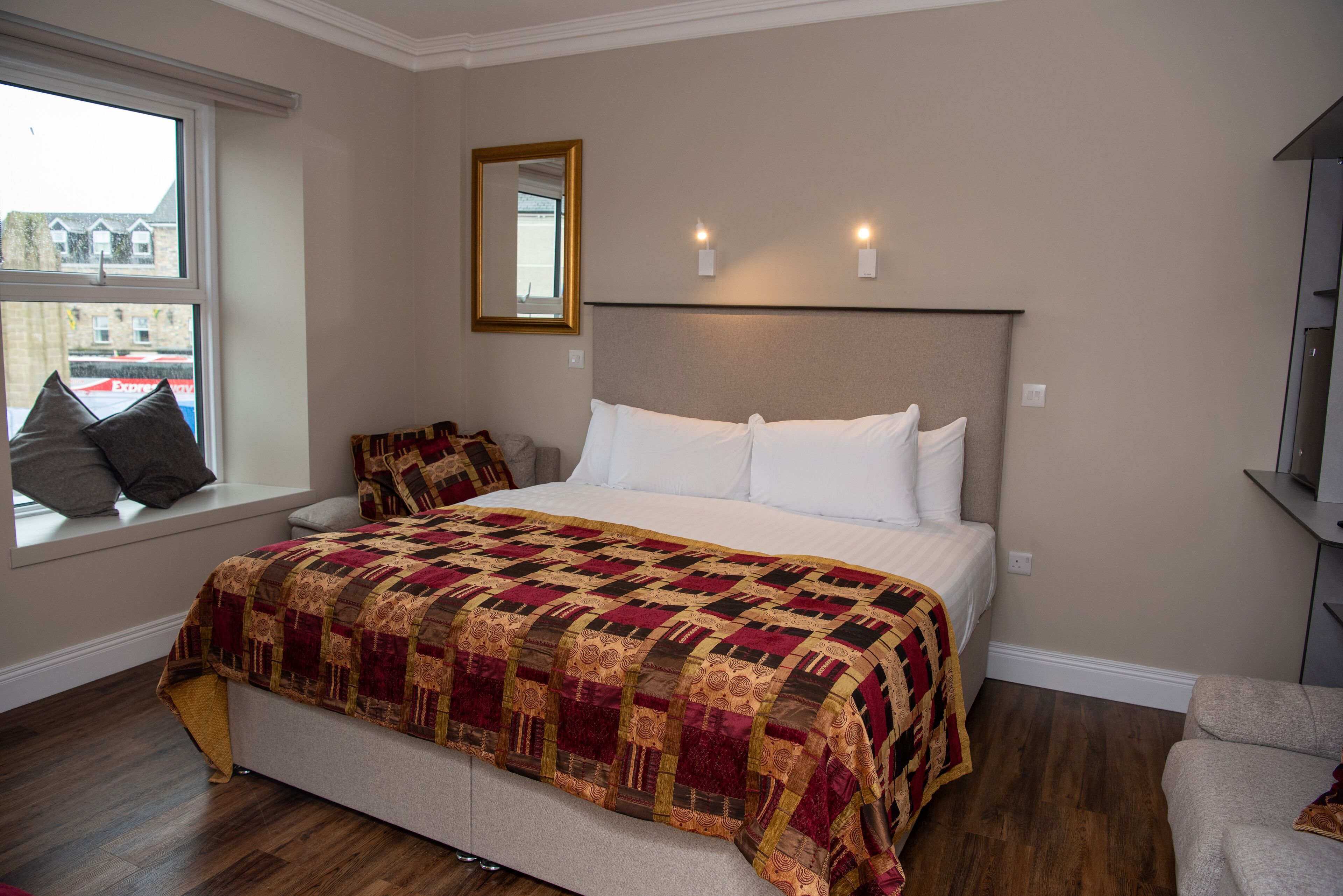 A bedroom with a large bed, a window with a view of buildings, a mirror, and a television on a stand.