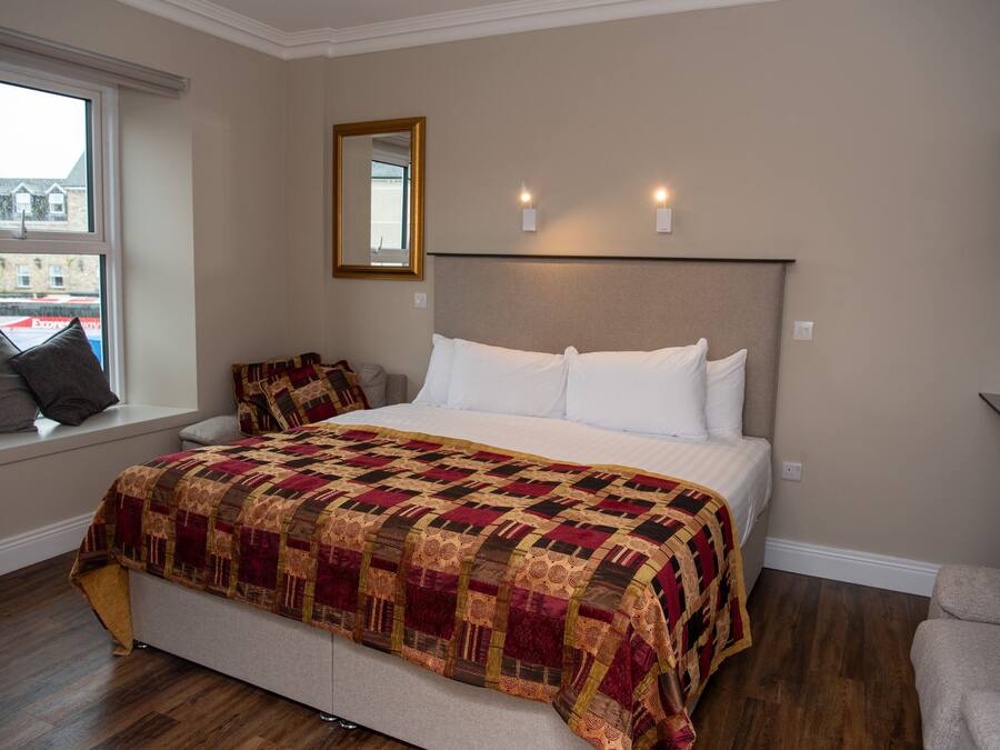 A bedroom with a large bed, a window with a view of buildings, a mirror, and a television on a stand.