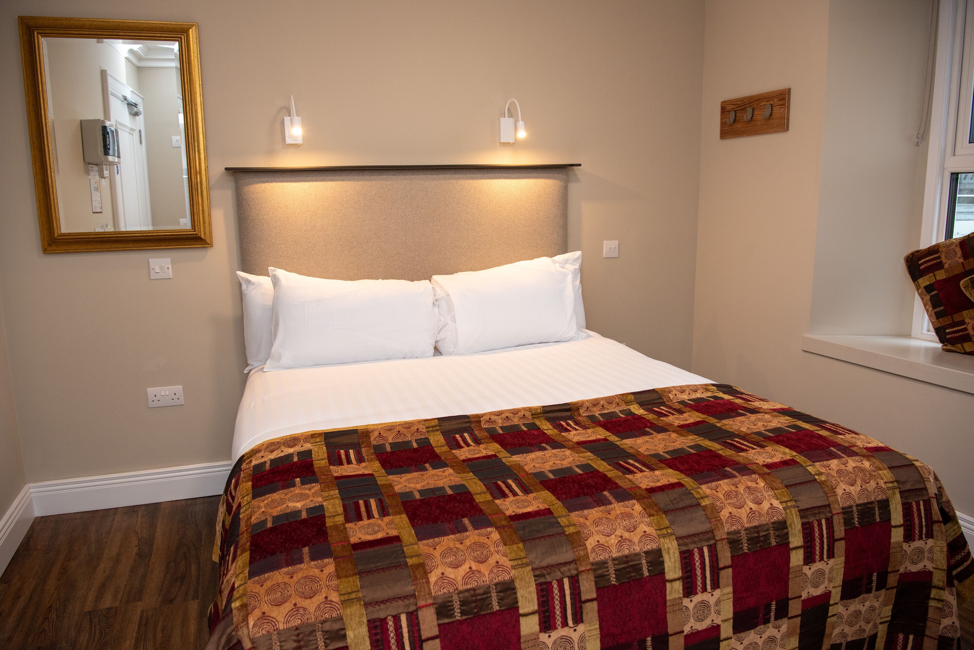 Una cama bien hecha con un edredón estampado, una lámpara de noche y un espejo en la pared.