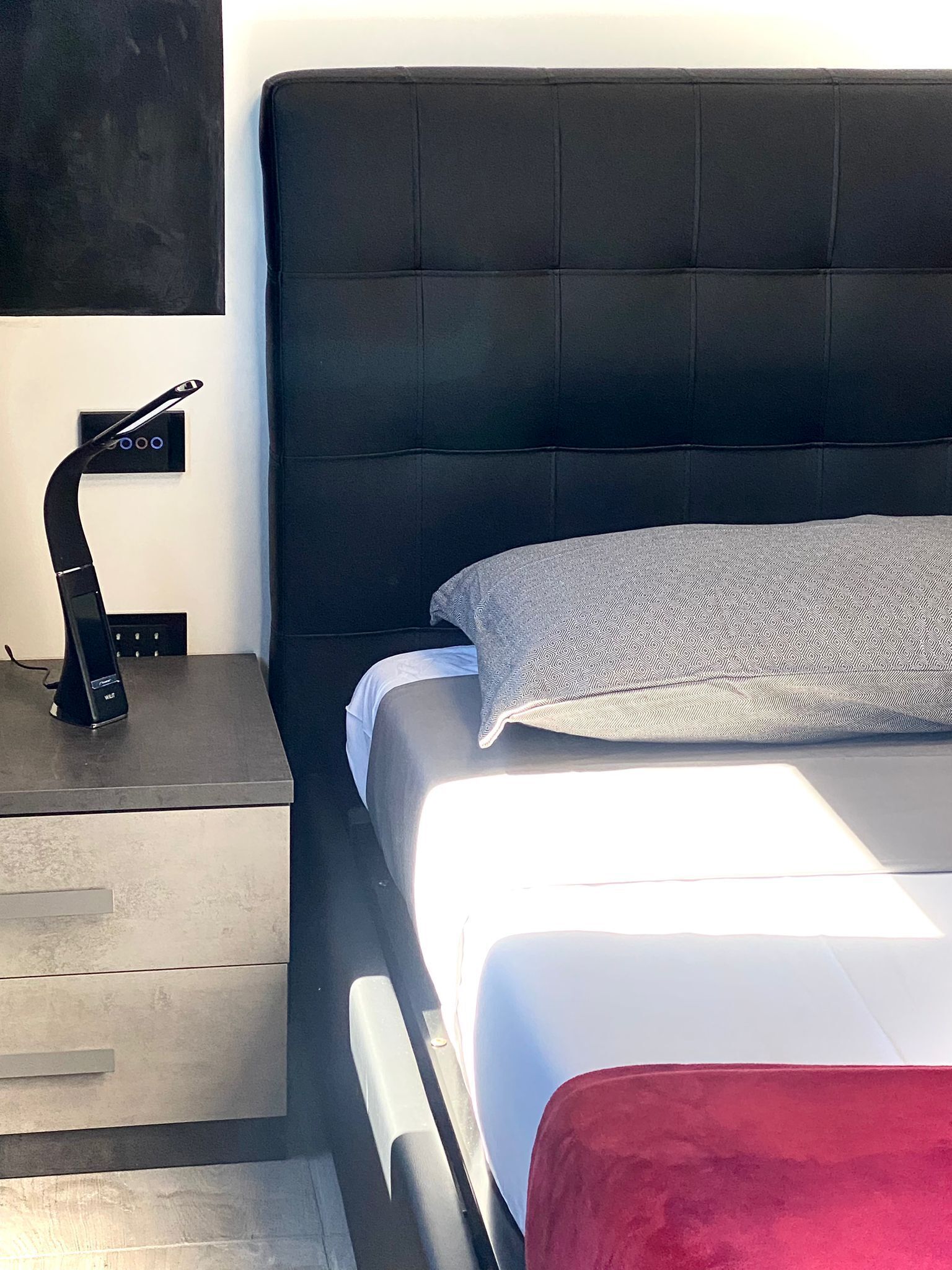 A modern hotel room with a large bed, a bedside table with a lamp, and a wall-mounted control panel.