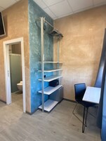 Un bagno piccolo e moderno con il water, un lavandino e uno spazio doccia. Si vede una scaffalatura con uno specchio e un porta asciugamani. Il pavimento è in piastrelle e c'è una porta che dà su un'altra stanza.