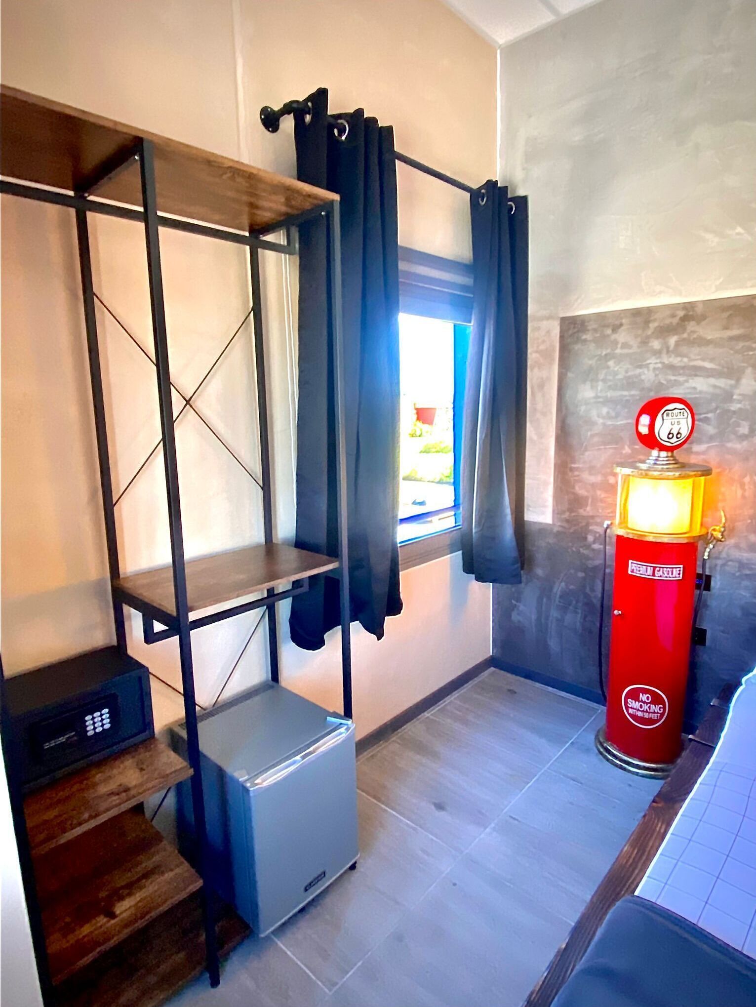 Quarto compacto com beliche, pequeno espaço de cozinha, janela com cortinas e placa vermelha de posto de gasolina.