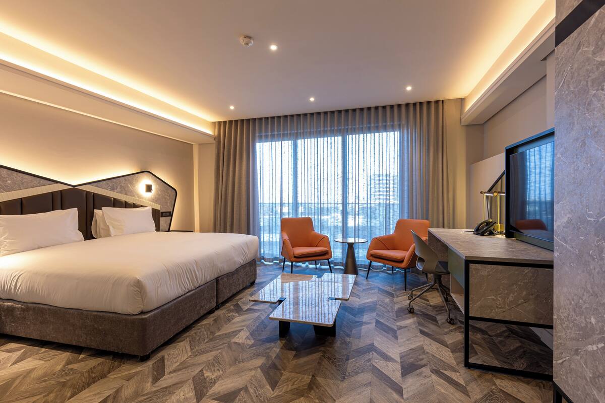 A modern hotel room with a large bed, two armchairs, a small table, and a desk with a chair.