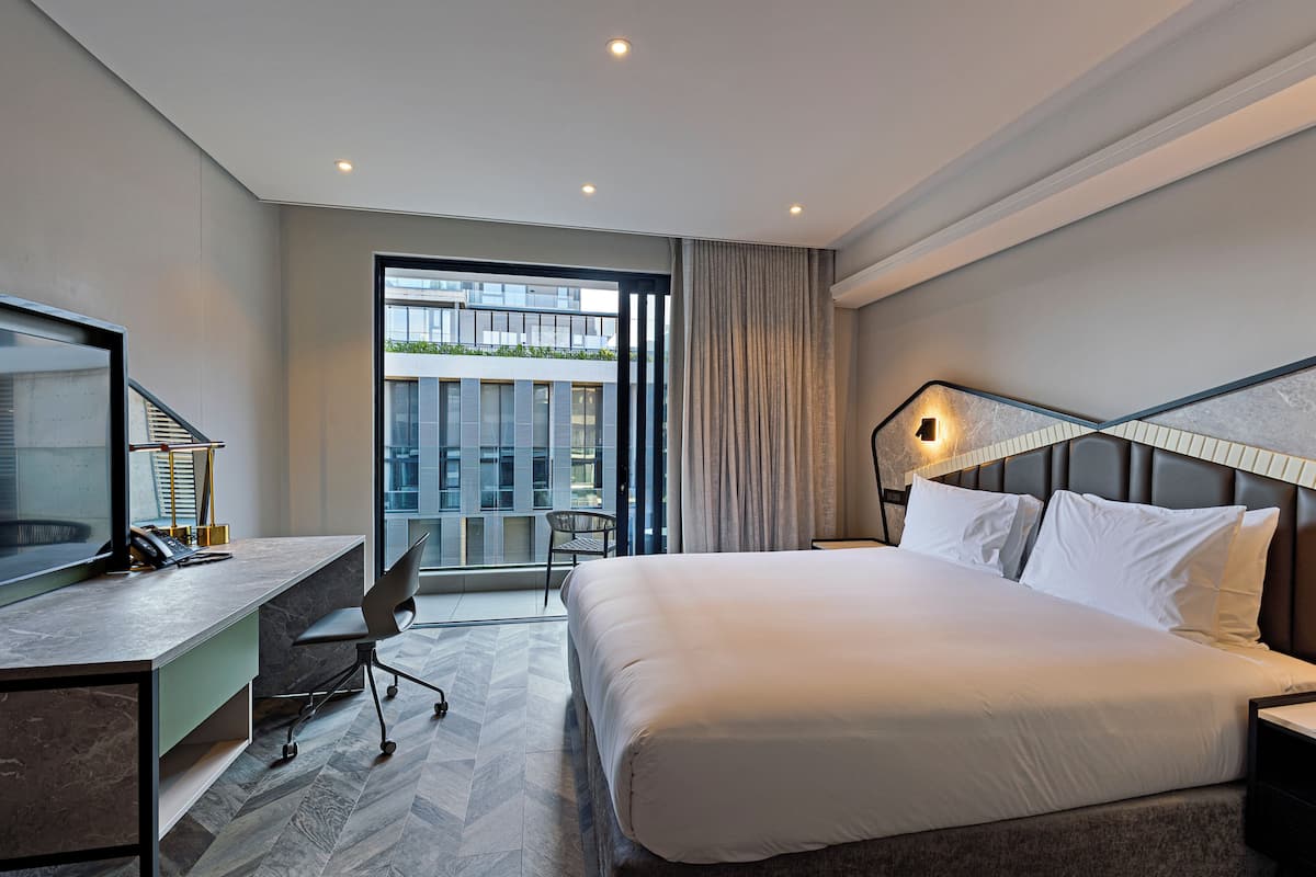A modern hotel room with a large bed, a desk with a chair, a television, and a view of the city through large windows.
