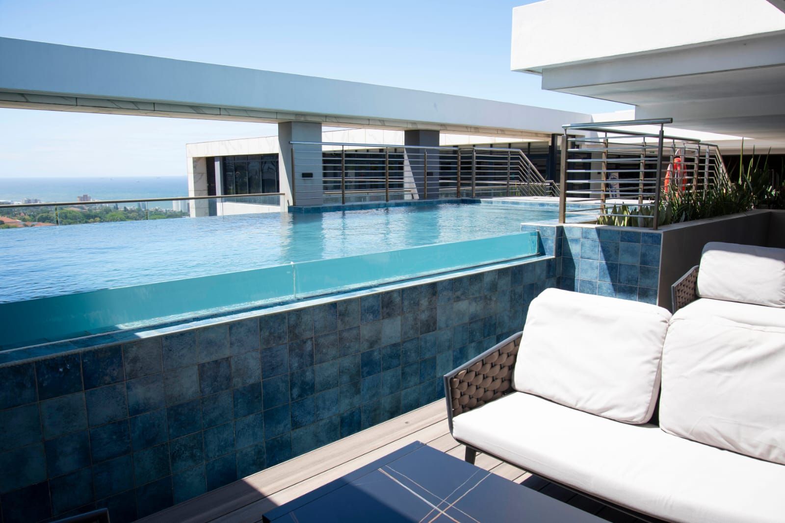 A modern outdoor pool area with a view of a cityscape, a white outdoor sofa, and a metal staircase.