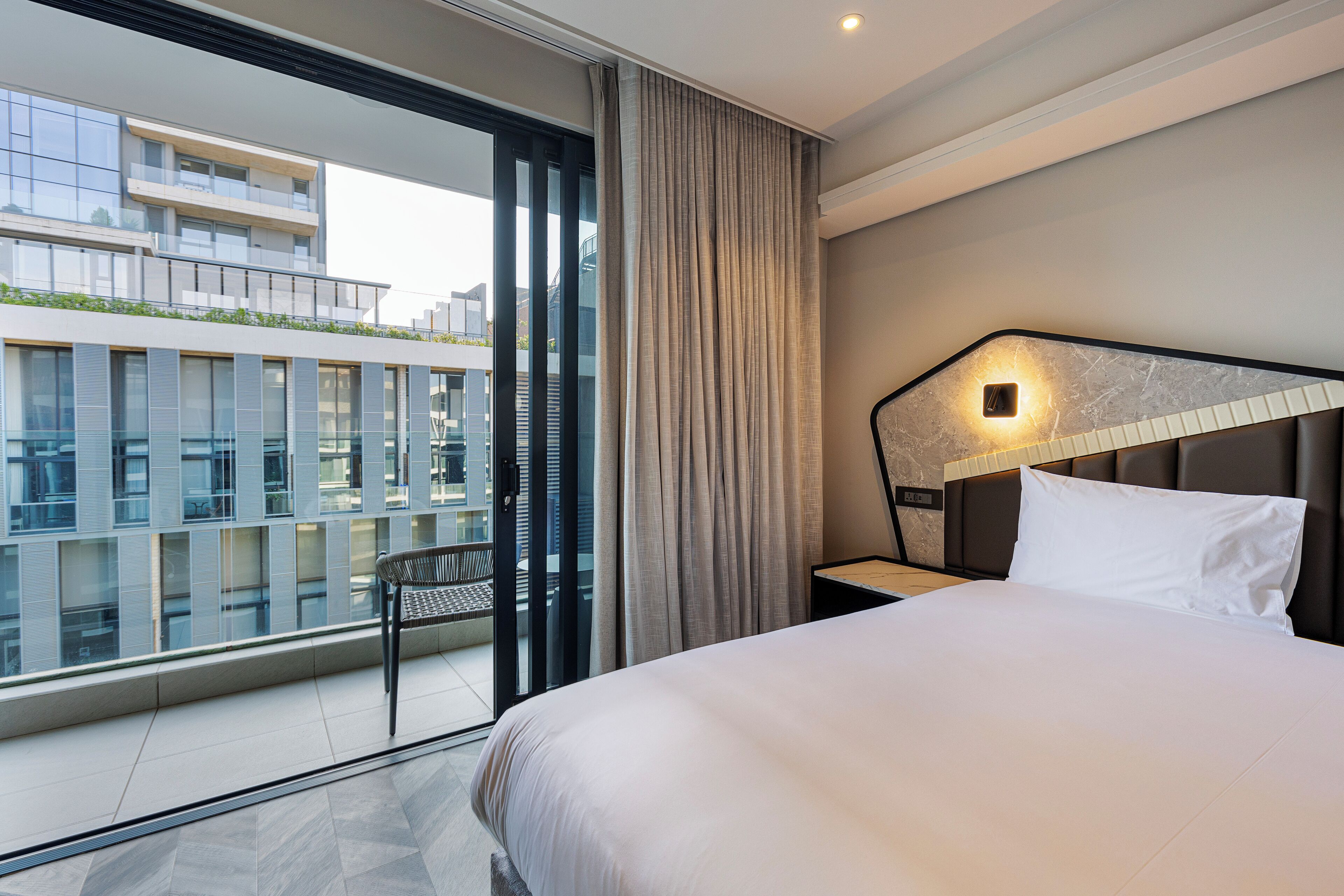 Een moderne hotelkamer met een groot bed, een nachtkastje, een stoel en uitzicht op de stad door een groot raam.