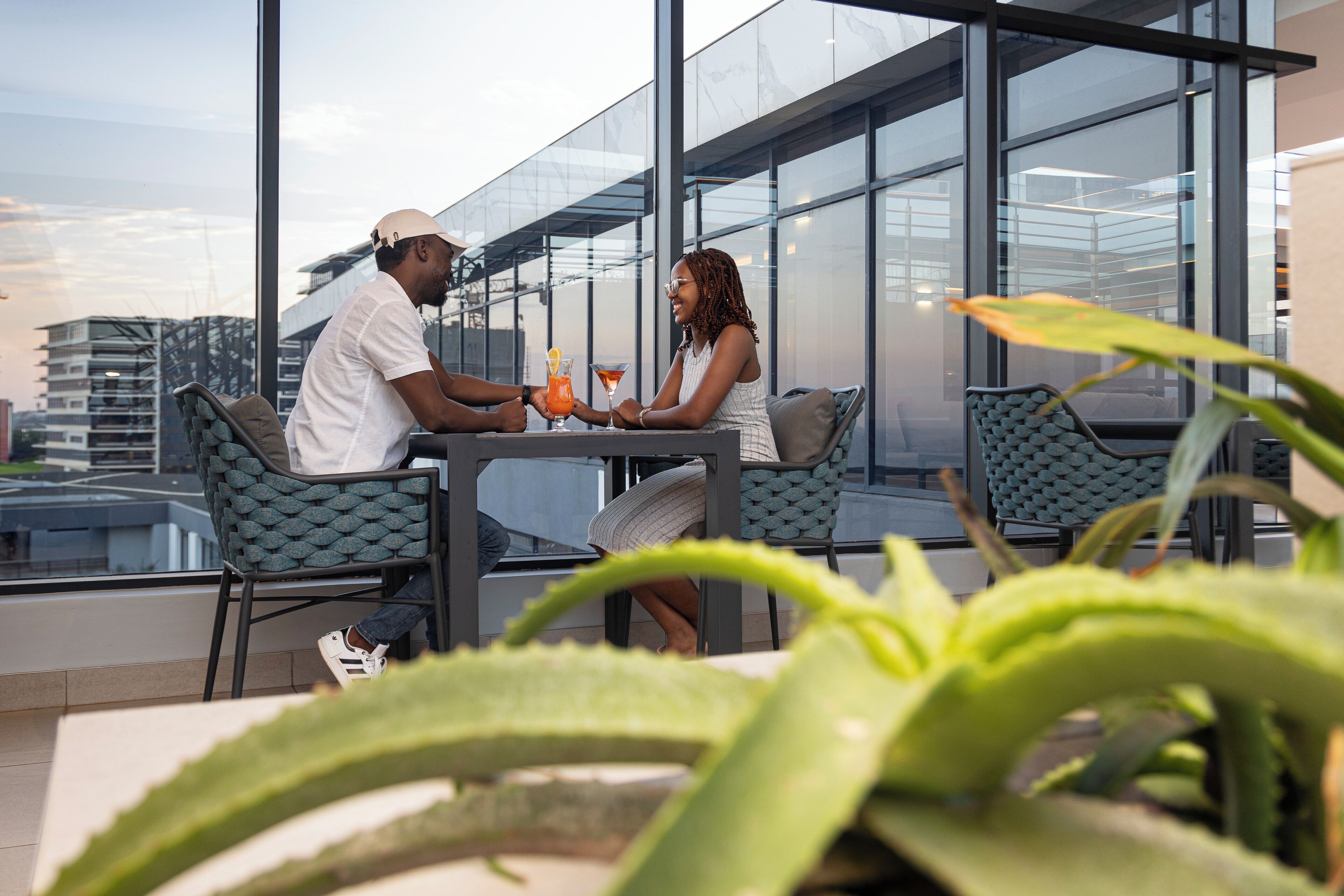 A modern outdoor seating area with a glass enclosure, featuring a table with drinks and two people conversing.