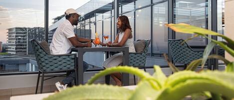 A modern outdoor seating area with a glass enclosure, featuring a table with drinks and two people conversing.