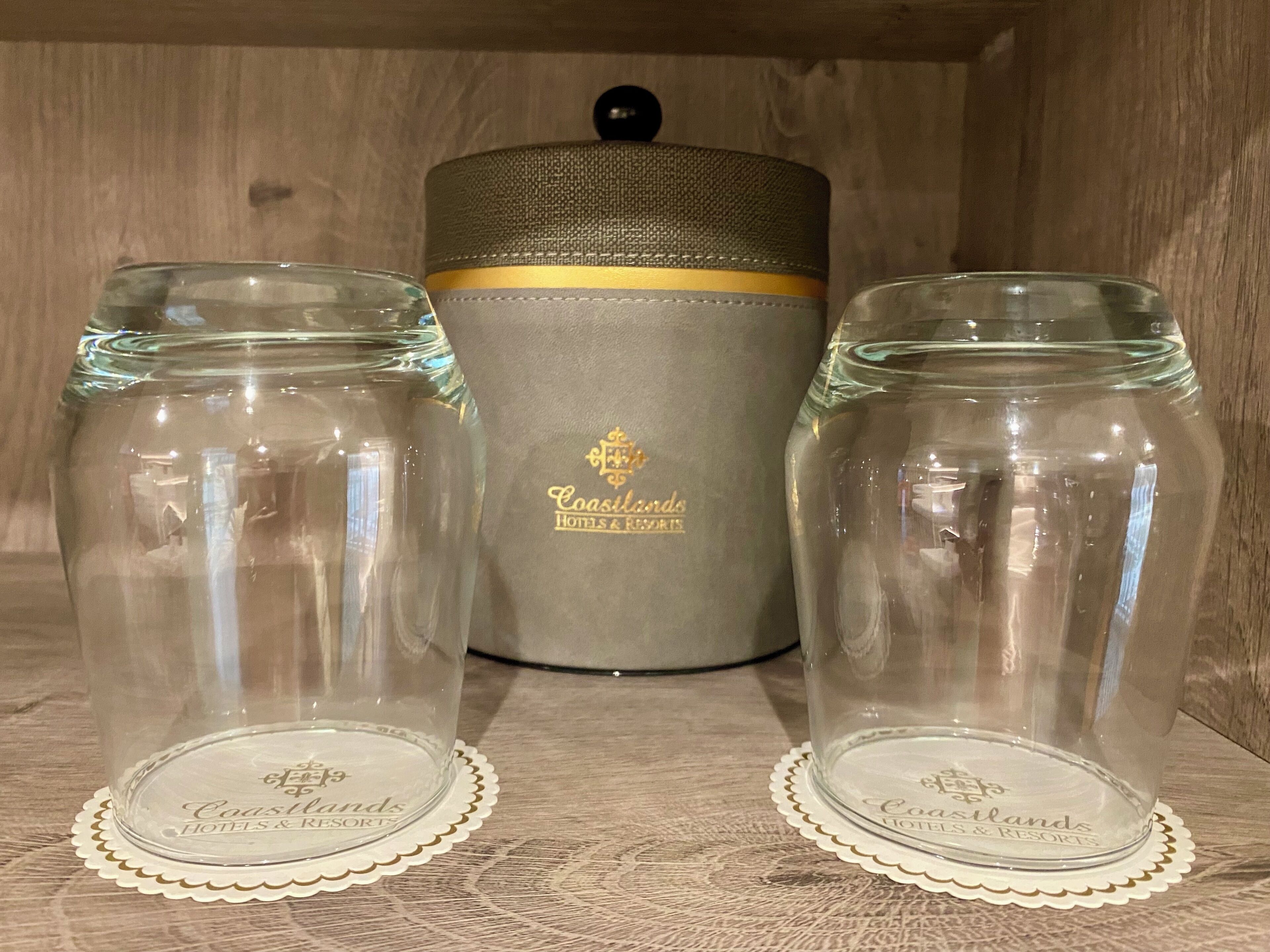 Two clear glass jars with lids, each on a white coaster with 'Coastlands Hotels & Resorts' branding, placed on a wooden surface with a grey container in the background.