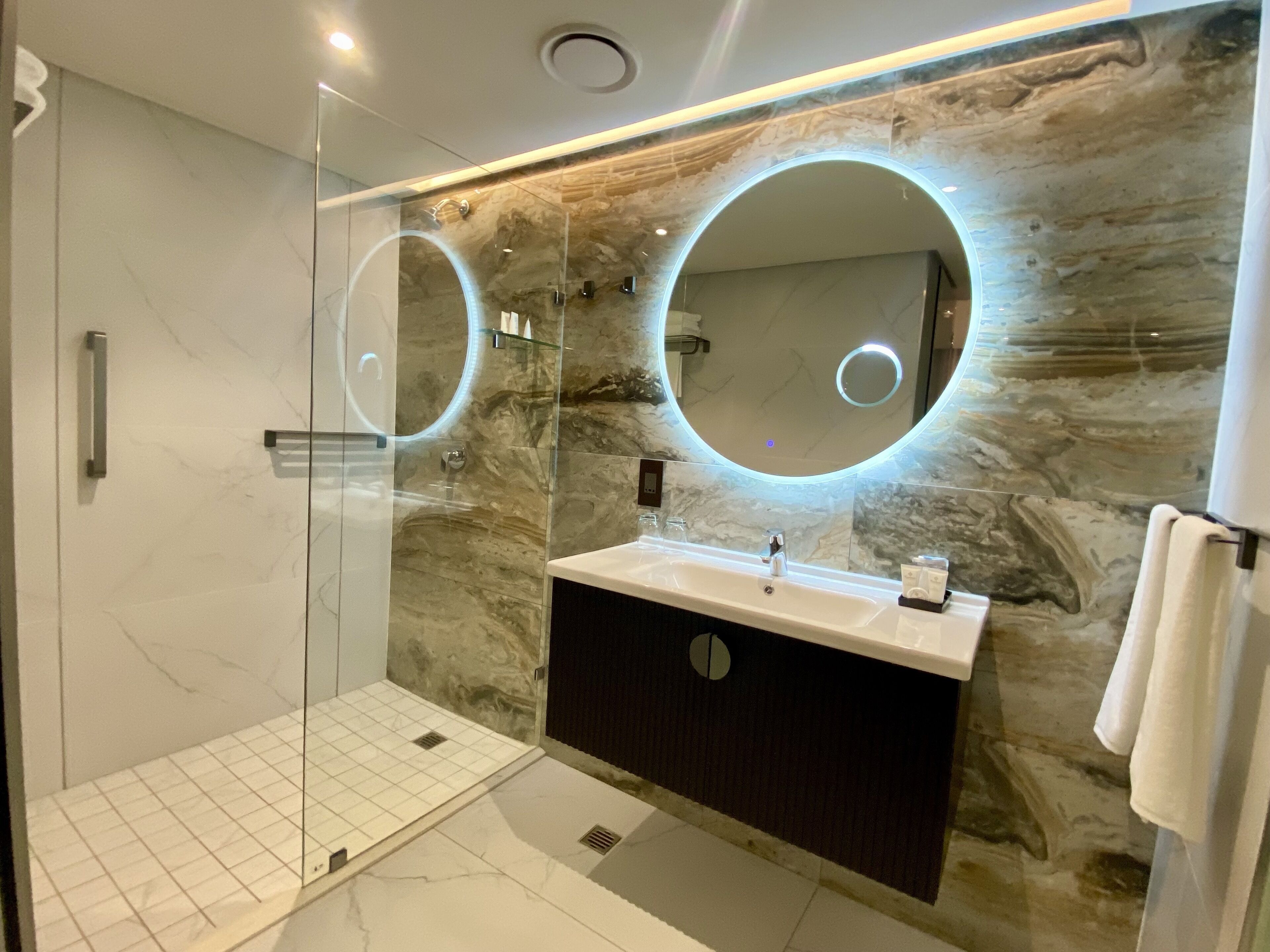 Een moderne badkamer met een douchecabine van glas, een wastafel met een marmeren spatwand en twee verlichte spiegels.