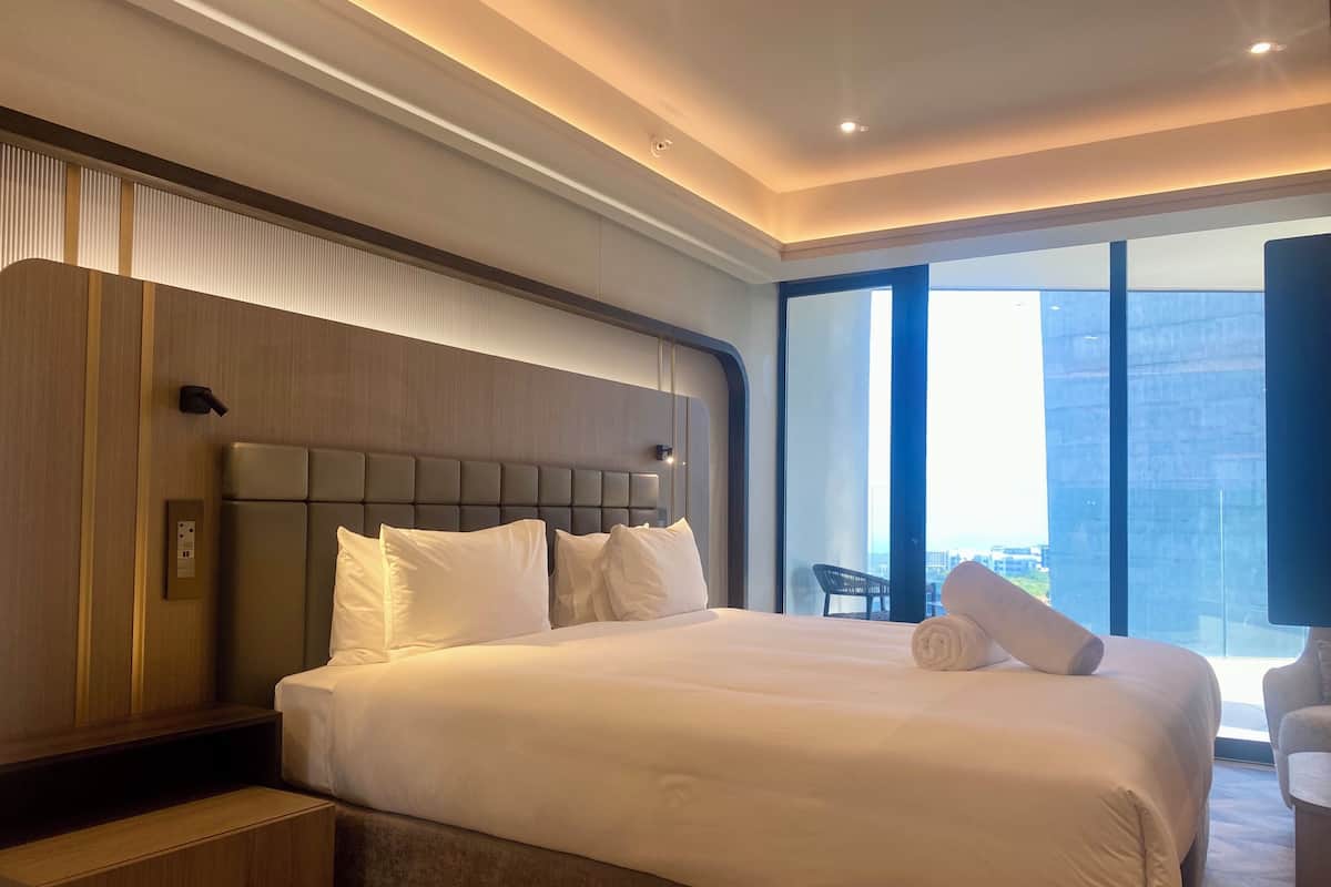 A modern hotel room with a large bed, a wooden headboard, a nightstand, and a view of the city through large windows.