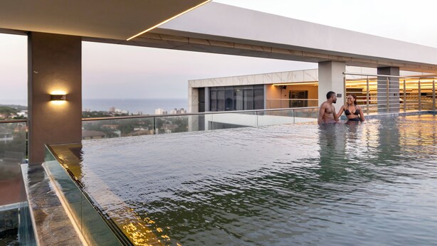 Una piscina infinita en la azotea con barandilla de cristal, un edificio moderno al fondo y vistas panorámicas de la ciudad.