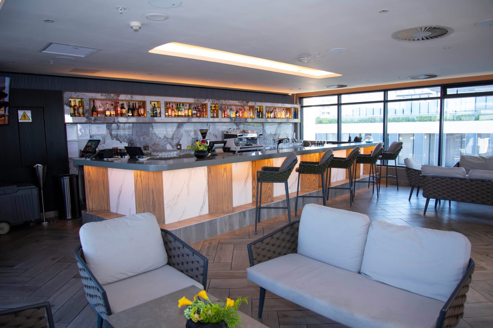 Uma área de bar moderna com bancada de mármore, banquetas e um espaço de estar com sofás e cadeiras.