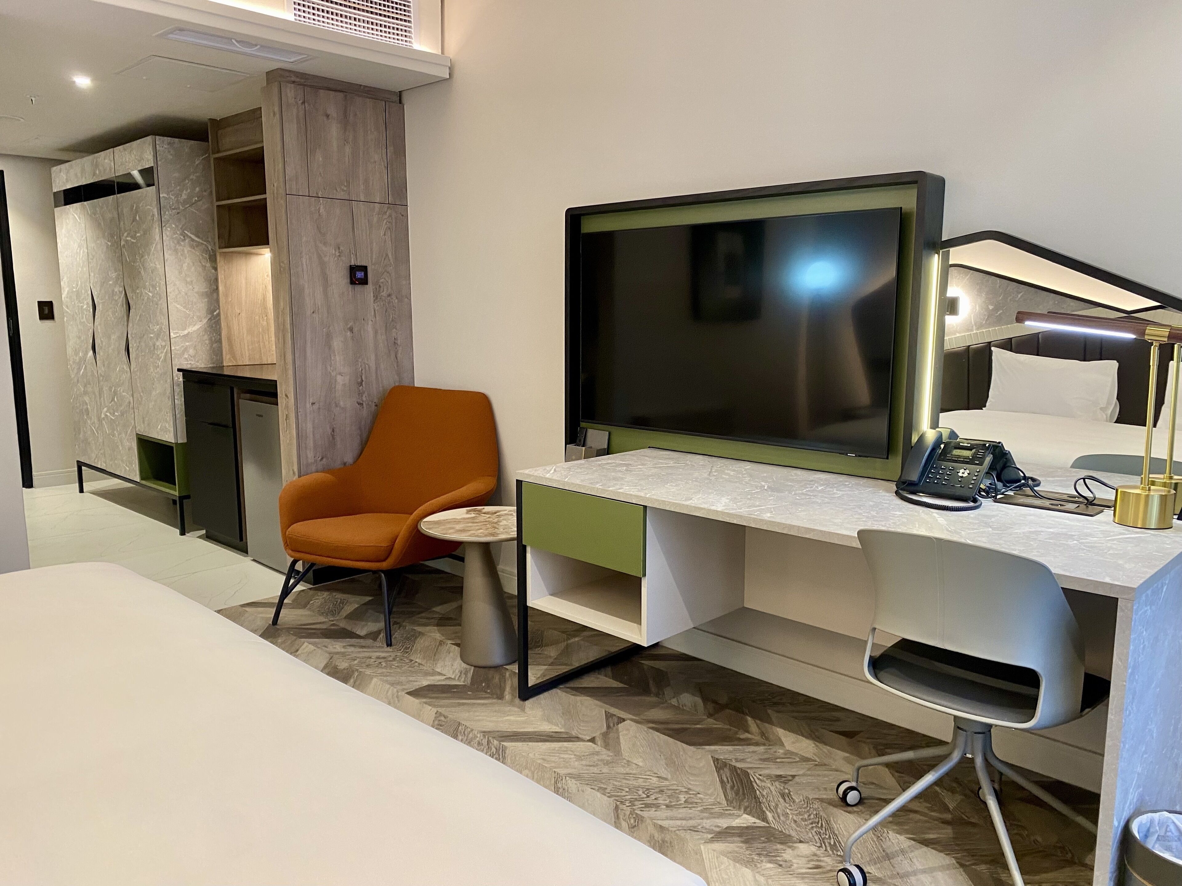 A modern hotel room with a large flat-screen TV, a desk with a telephone, an orange chair, and a bed with white bedding.