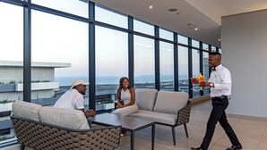 Un espacio interior moderno con ventanas amplias, un conjunto de asientos y una persona caminando con una bebida.