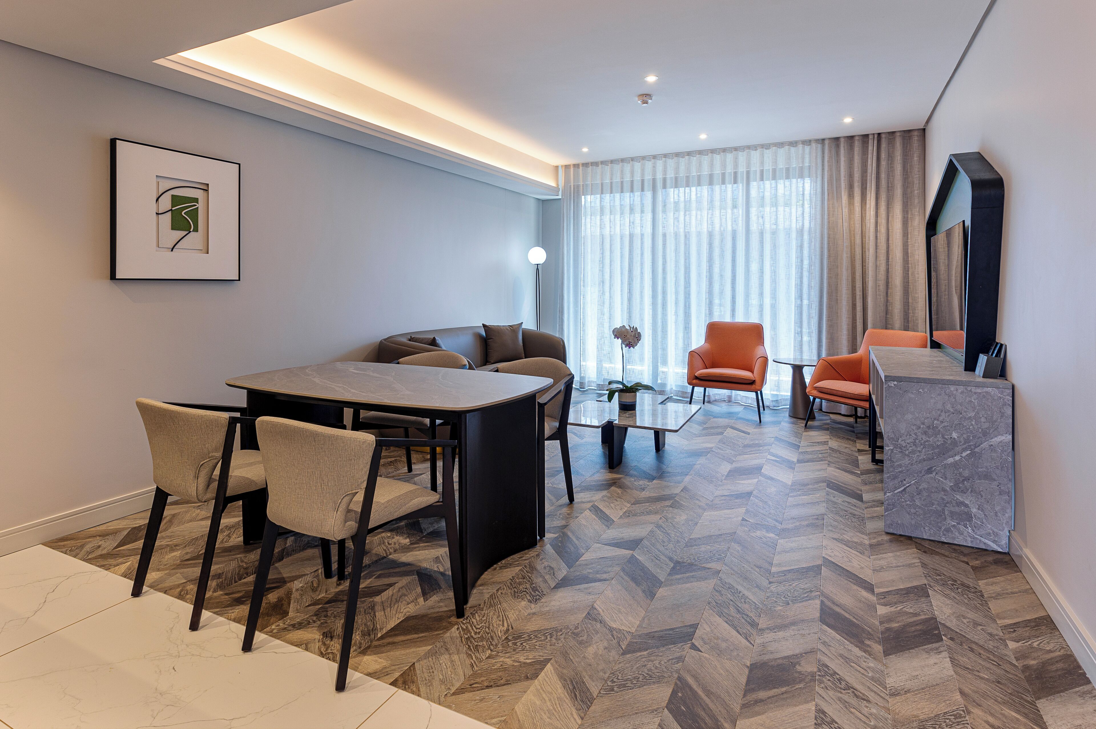 A modern hotel room with a dining area, a sofa, two armchairs, a coffee table, and a large mirror.