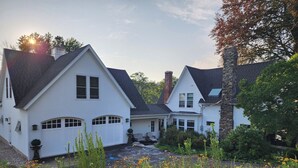 Une maison blanche à deux étages, dotée d’une cheminée en pierre, entourée d’un jardin où poussent diverses plantes et fleurs.