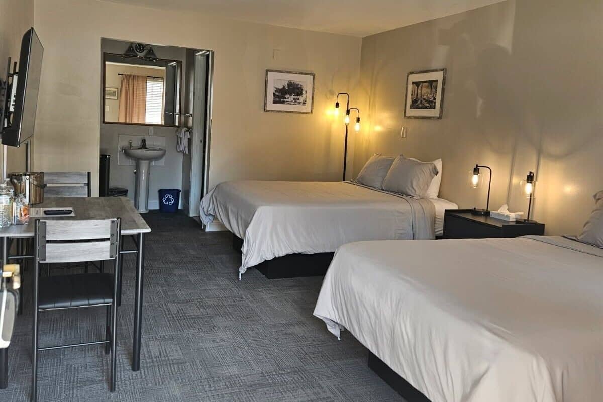 A hotel room with two beds, a desk, a chair, a sink, and two wall-mounted lamps.