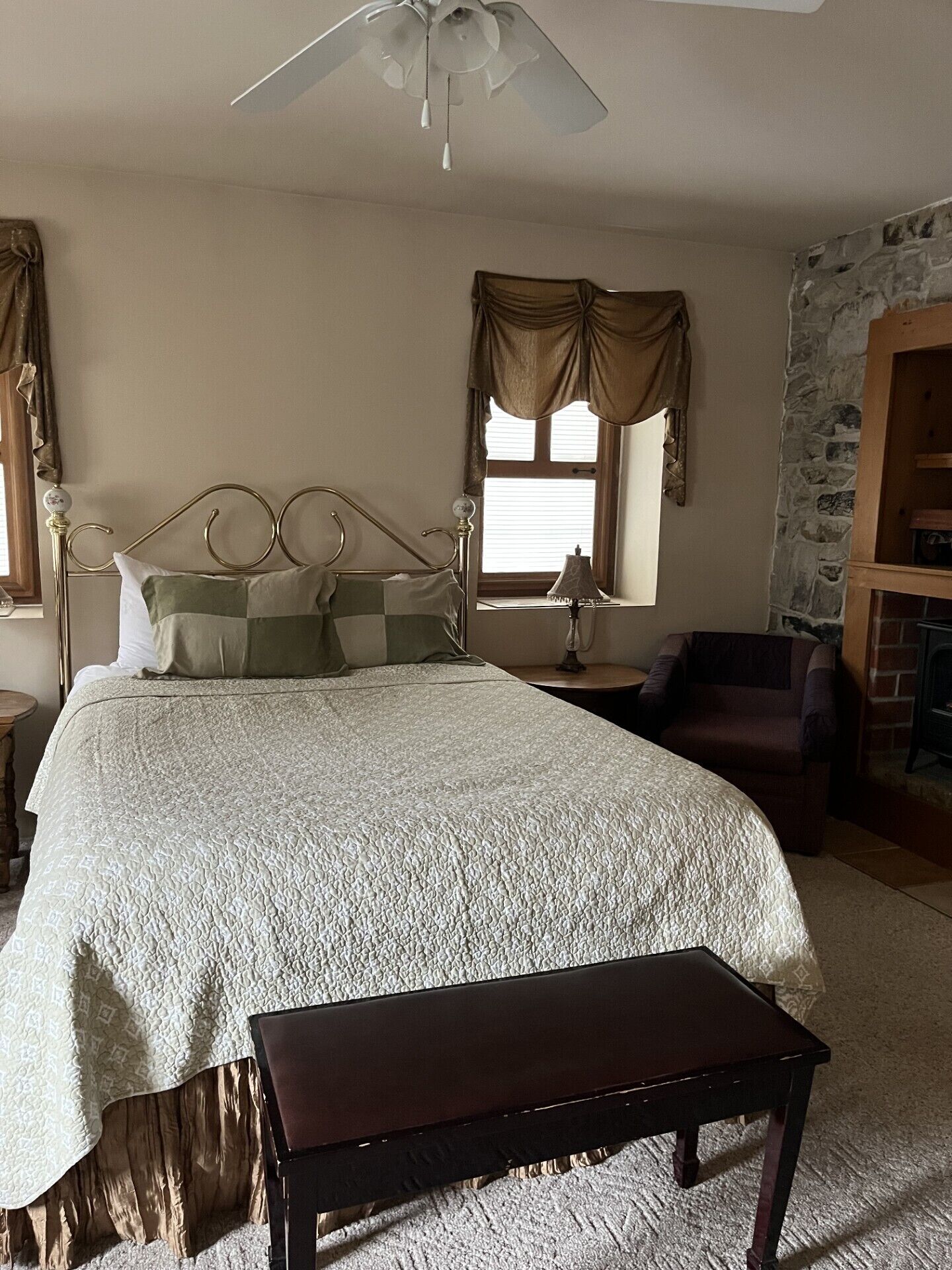 Un dormitorio con cama, mesita de noche, lámpara, silla, banco, ventilador de techo, ventana con cortinas y pared de piedra.