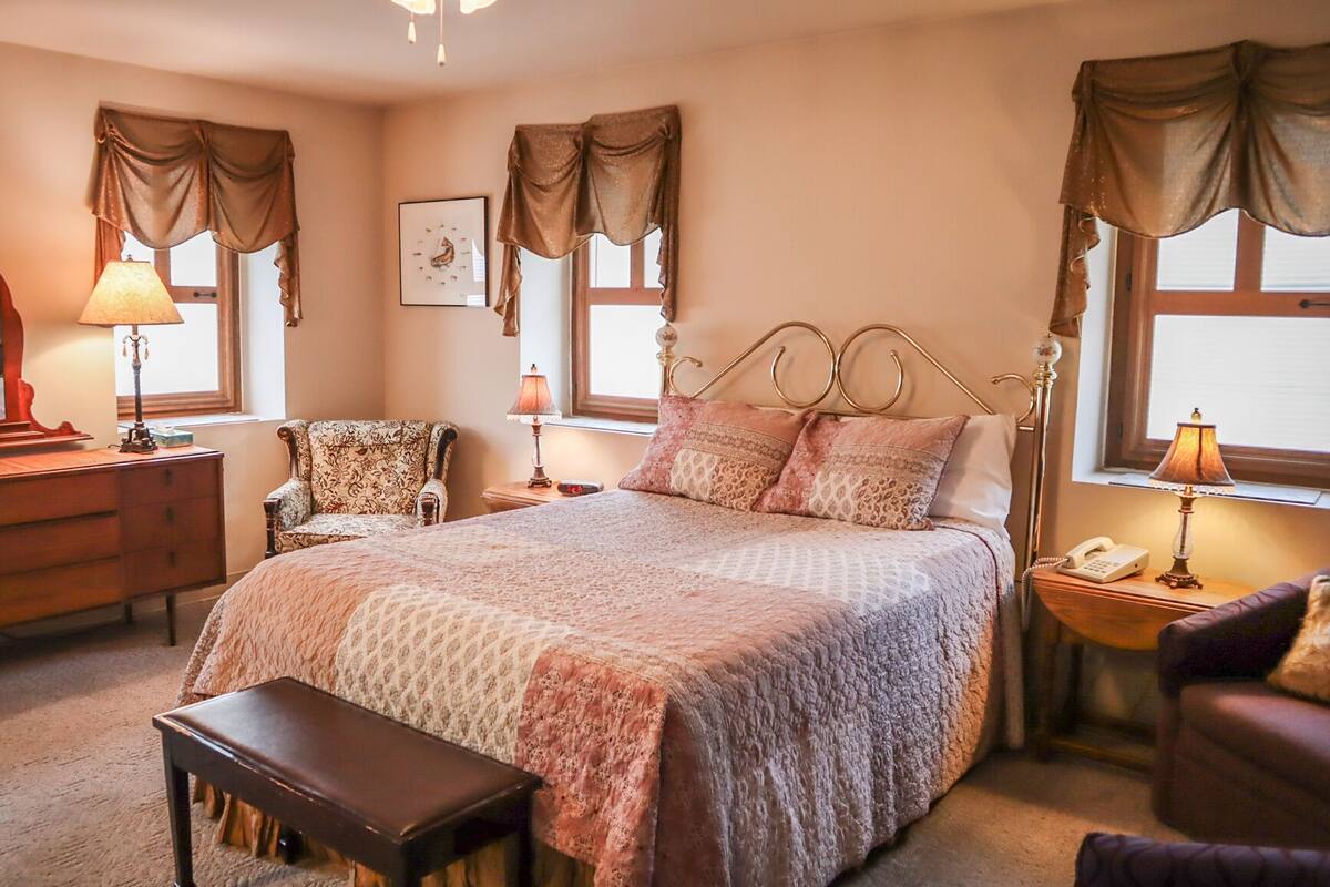A bedroom with a large bed, a wooden dresser, a chair, a bench, and two bedside tables with lamps.