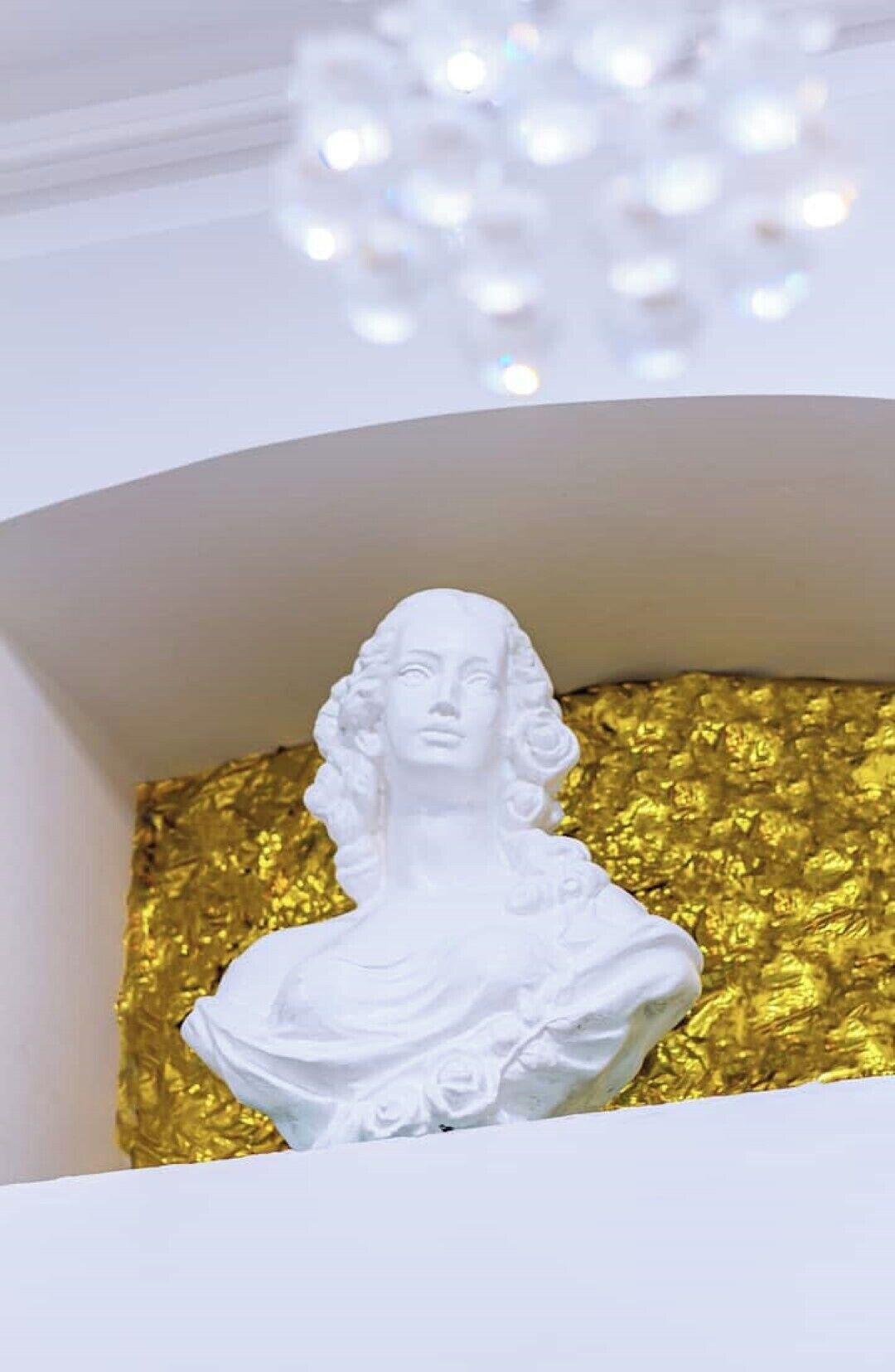 A white bust sculpture in a white alcove with a golden textured backdrop.