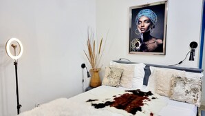 Un dormitorio con cama, lámpara de pie, jarro con plantas secas y un cuadro en la pared.