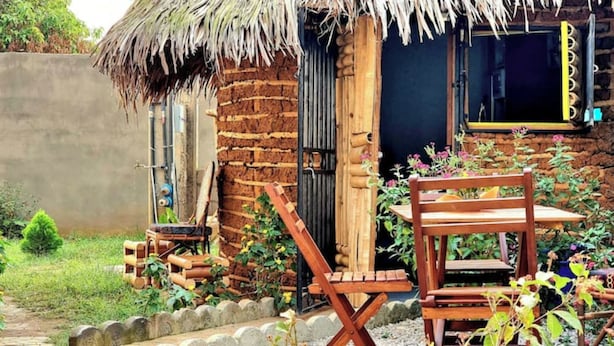 Una casa de madera rústica con techo de paja, un banco de madera y un pequeño jardín con plantas y flores.