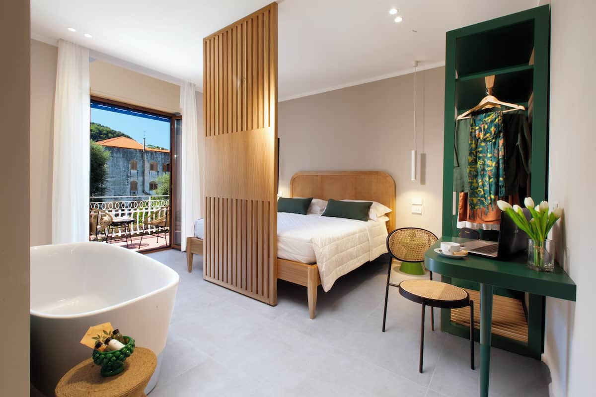 Ein modernes Hotelzimmer mit einer großen Badewanne, einem Bett mit Holz-Kopfteil, einem Schreibtisch mit Laptop und Blick auf einen Balkon mit Gartenmöbeln.
