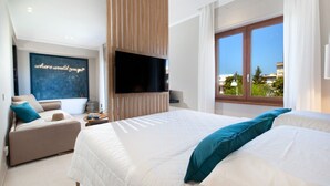 A modern hotel room with a large bed, a sofa, a TV mounted on the wall, and a view of the city through the window. - Estate4home - Suites 21 (Sorrento)