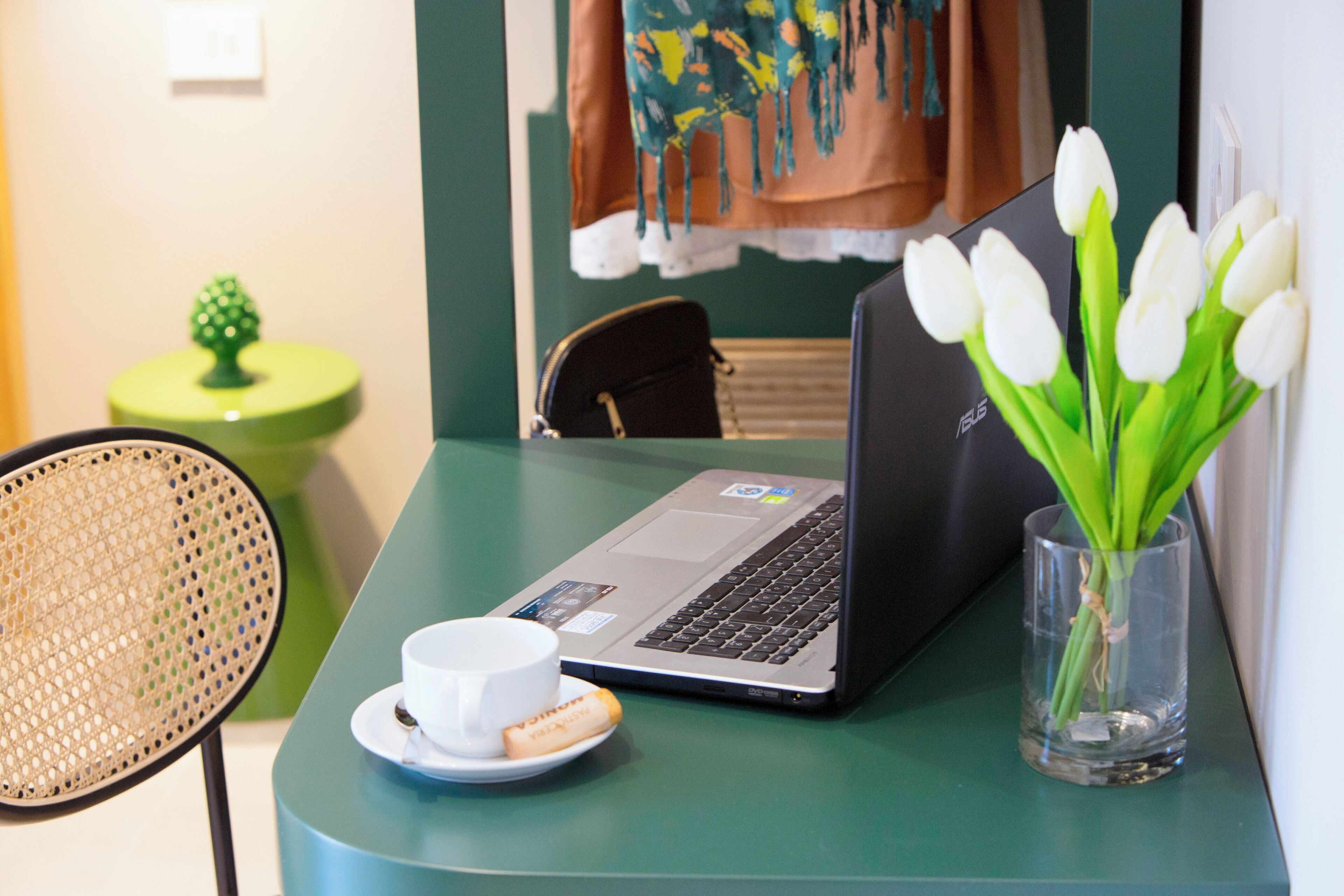Un ordinateur portable est posé sur une table, à côté d’une tasse et d’une assiette de biscuits. On aperçoit également une chaise, une petite table ornée d’un objet décoratif et un vase contenant des fleurs blanches.