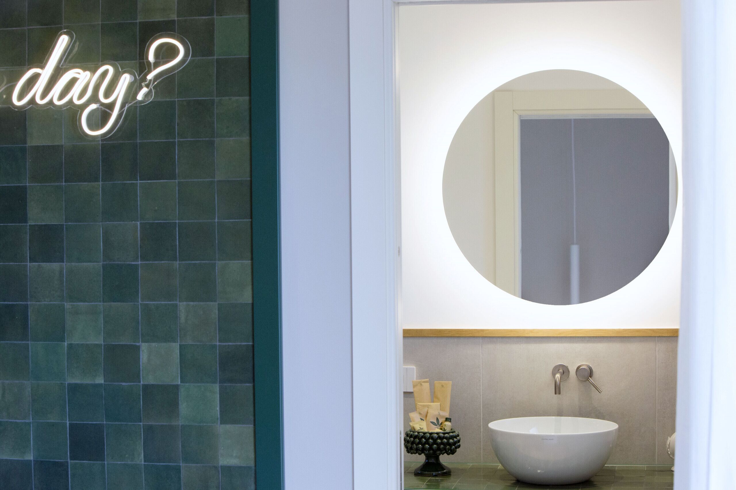 Un baño con un espejo redondo, un lavabo blanco y un letrero de neón que dice '¿día?'.