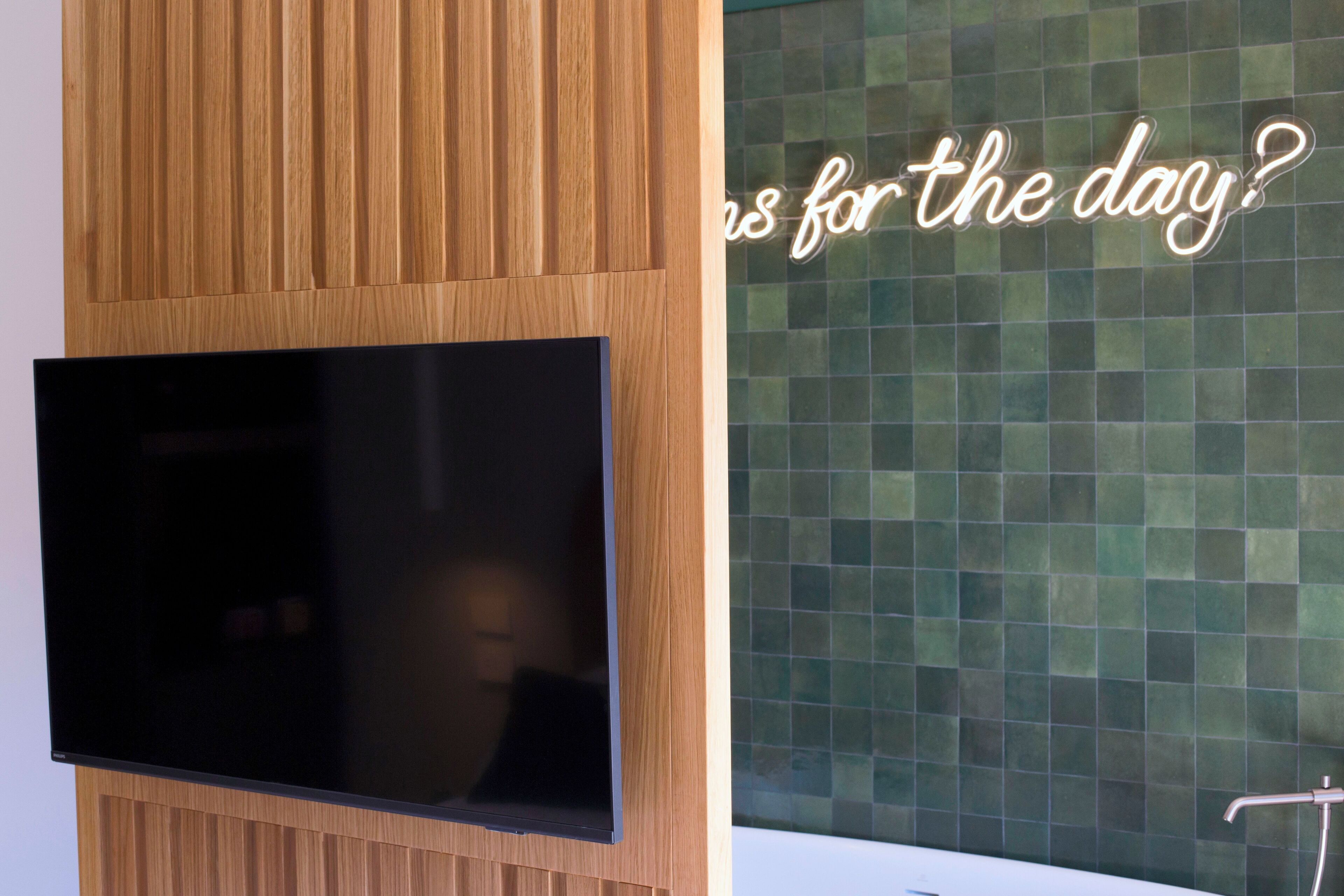 Un baño moderno con pared de paneles de madera, un televisor de pantalla plana montado en ella, y pared de mosaicos verdes con un letrero de neón que dice '¿Es esto para el día?'.