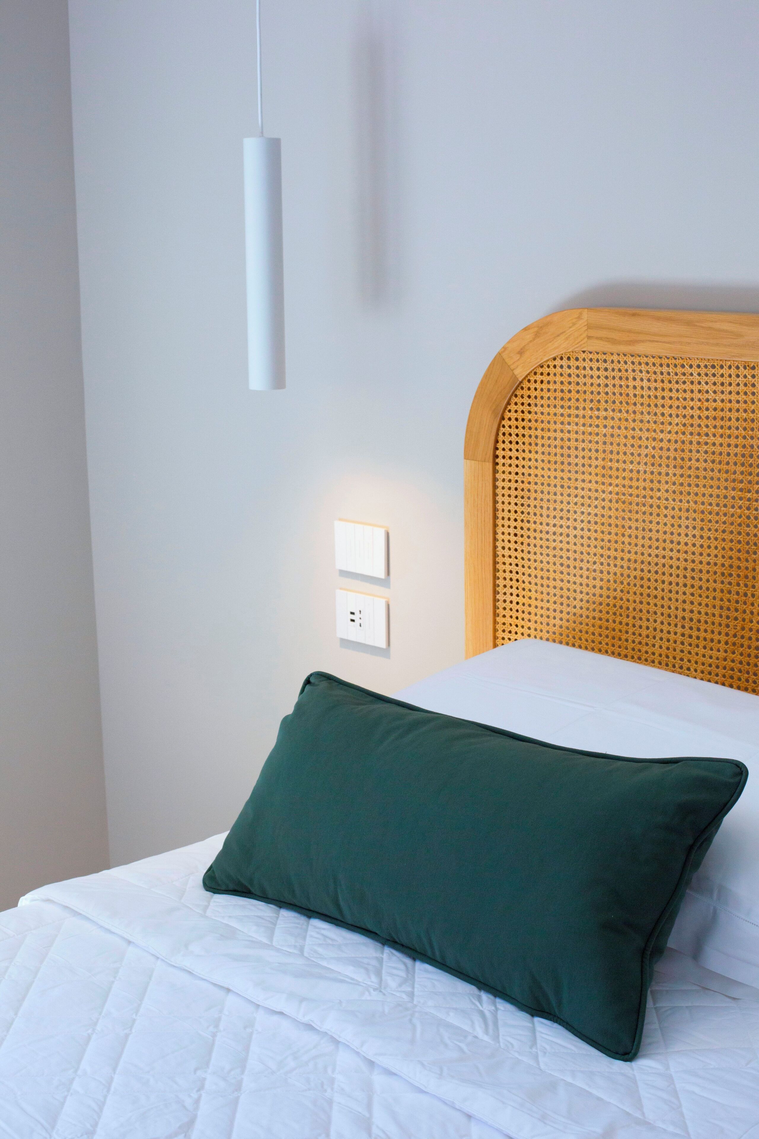 Een net opgemaakt bed met een groen kussen, een wit hoofdbord en een eenvoudig wandlampje.
