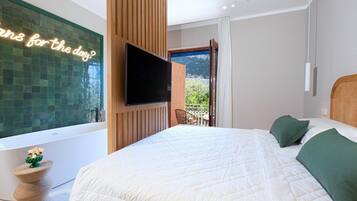 Un dormitorio moderno con una cama grande, un televisor de pantalla plana montado en un panel de madera, una bañera y un balcón con vistas a la vegetación.