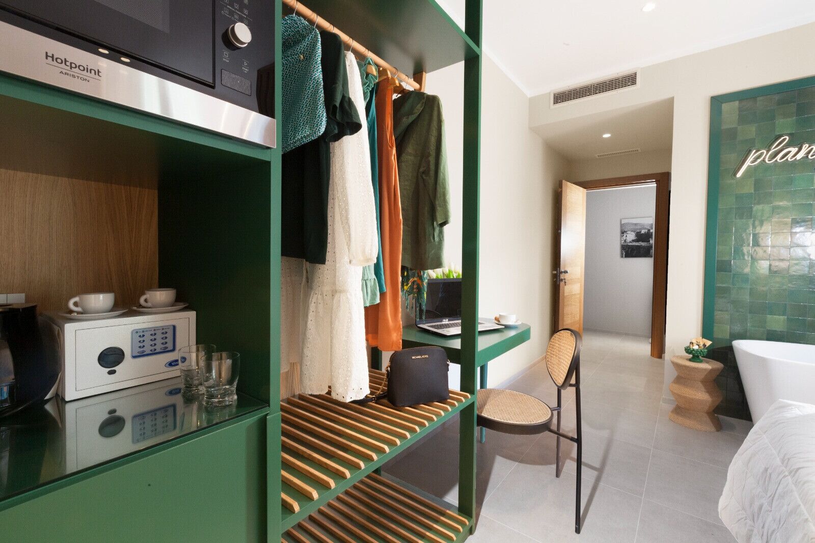 A hotel room with a kitchenette, a bed, a desk with a laptop, a chair, and a bathroom with a green tiled wall.