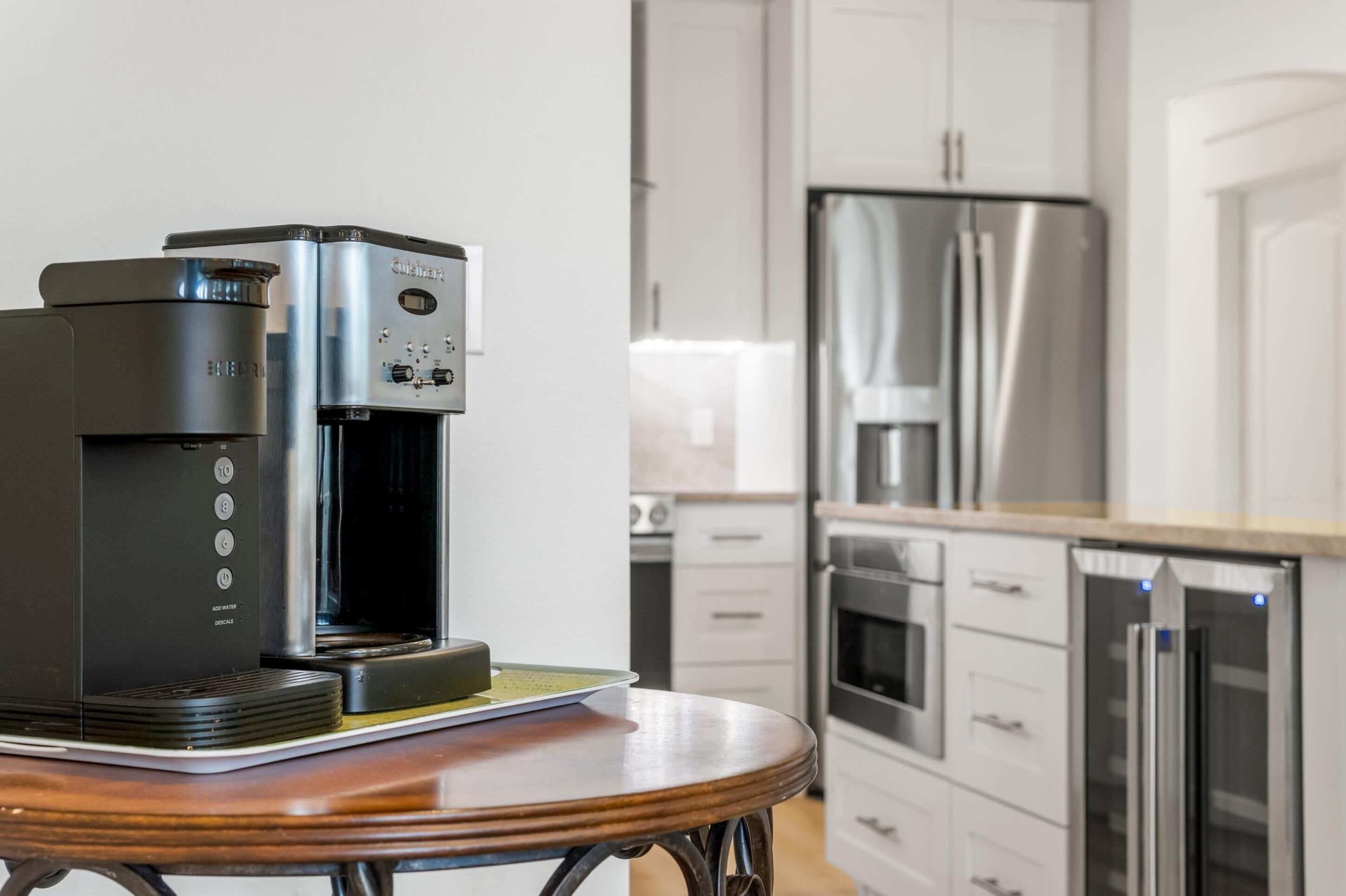 Una cocina moderna con una cafetera sobre una mesa, electrodomésticos de acero inoxidable y armarios blancos.