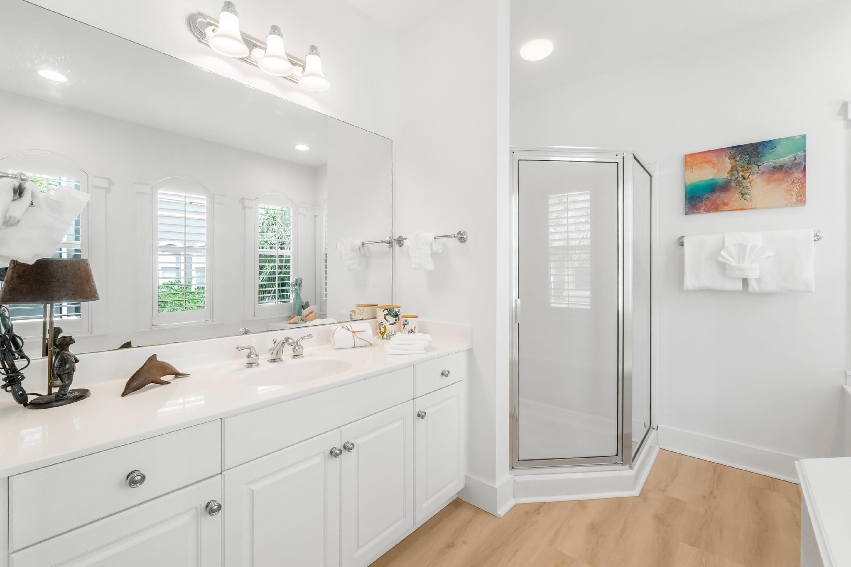 Un baño moderno con un espejo grande, muebles blancos, ducha con puertas de vidrio y un cuadro abstracto de colores en la pared.