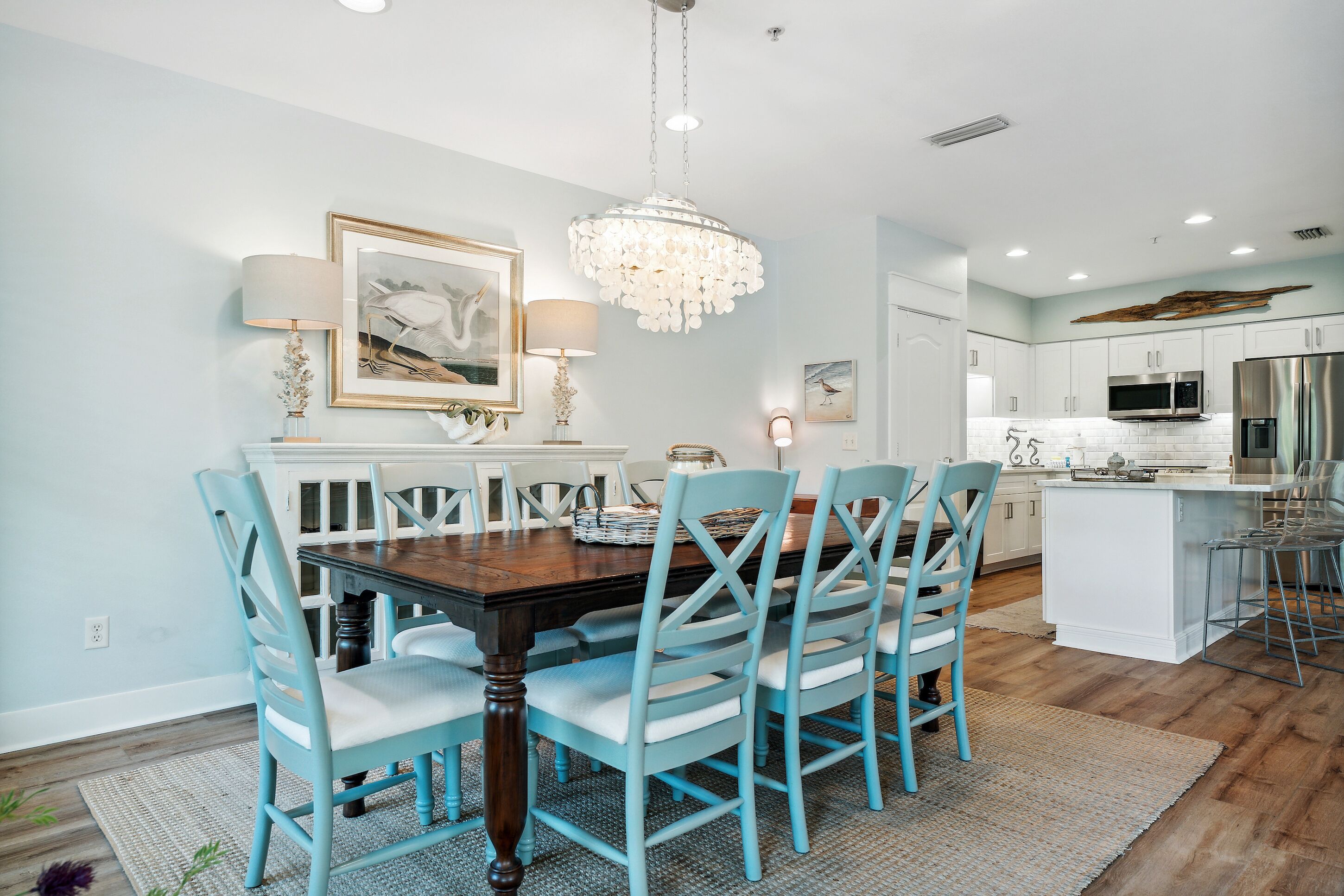 A dining area with a wooden table and blue chairs, a chandelier, and a kitchen with white cabinets and stainless steel appliances.